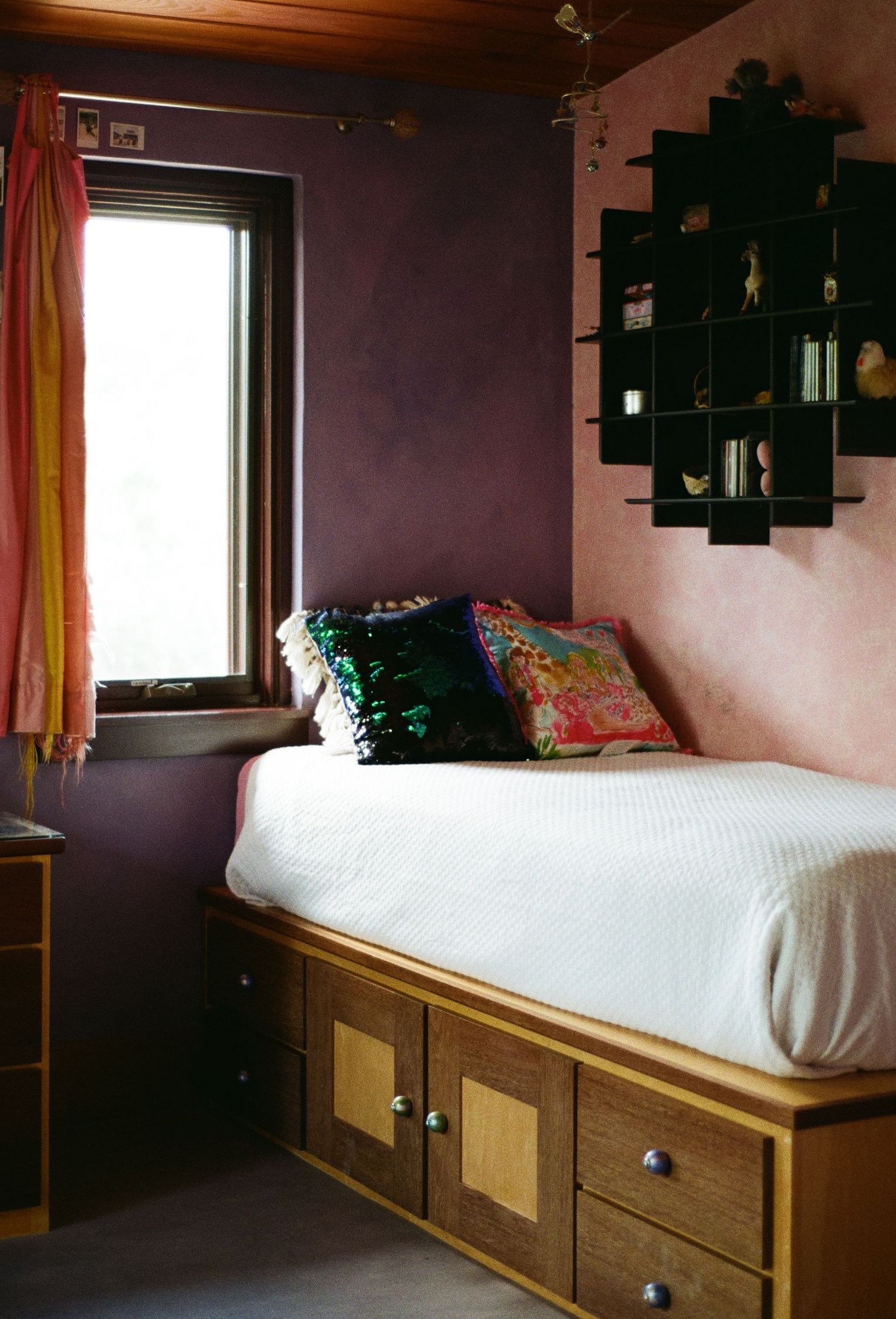 Cozy bedroom with platform bed, storage drawers, purple walls, colorful pillows, and decorative wall-mounted display shelf.