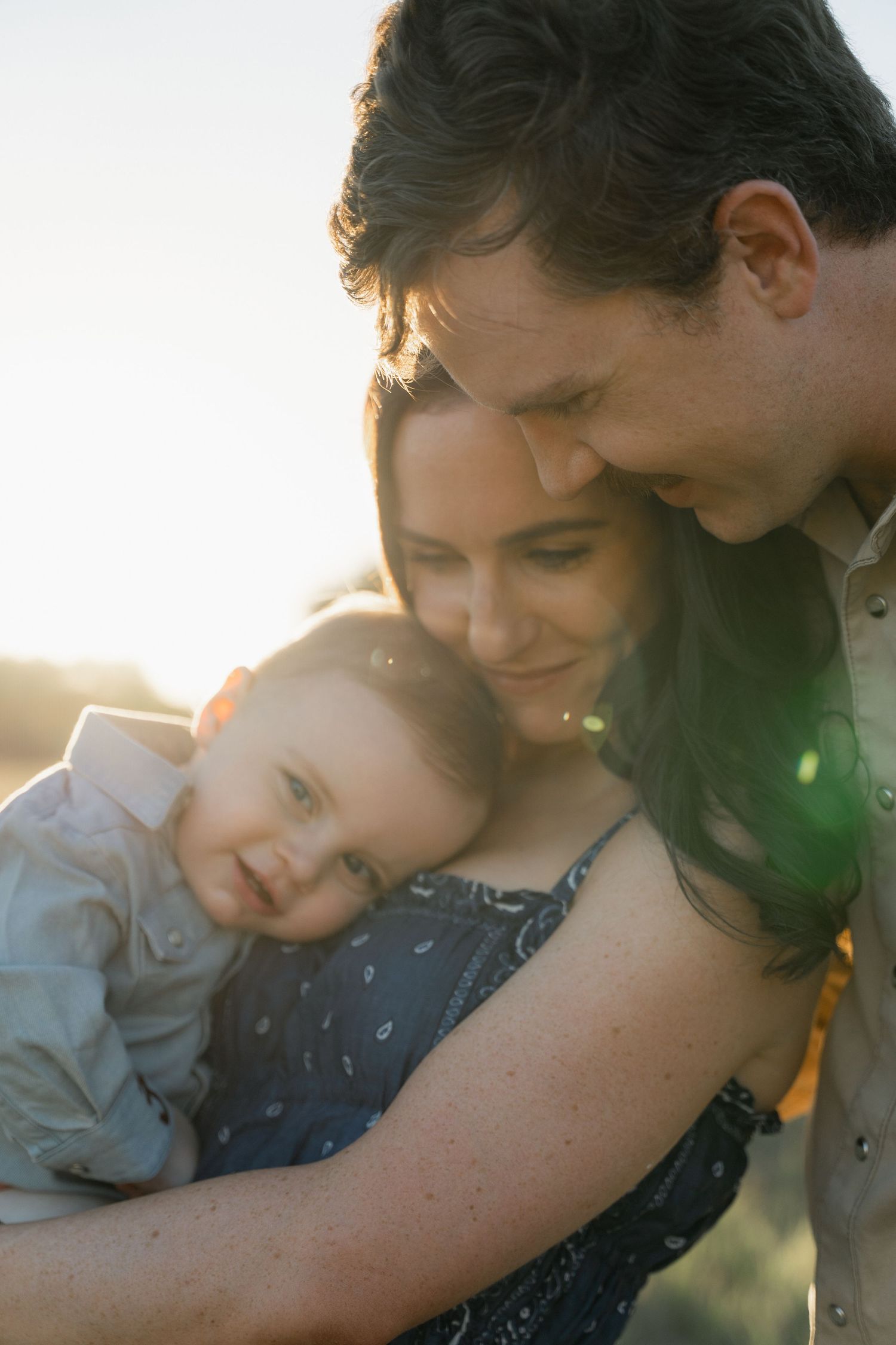 Parents holding smiling baby in golden sunset light during family portrait session.
