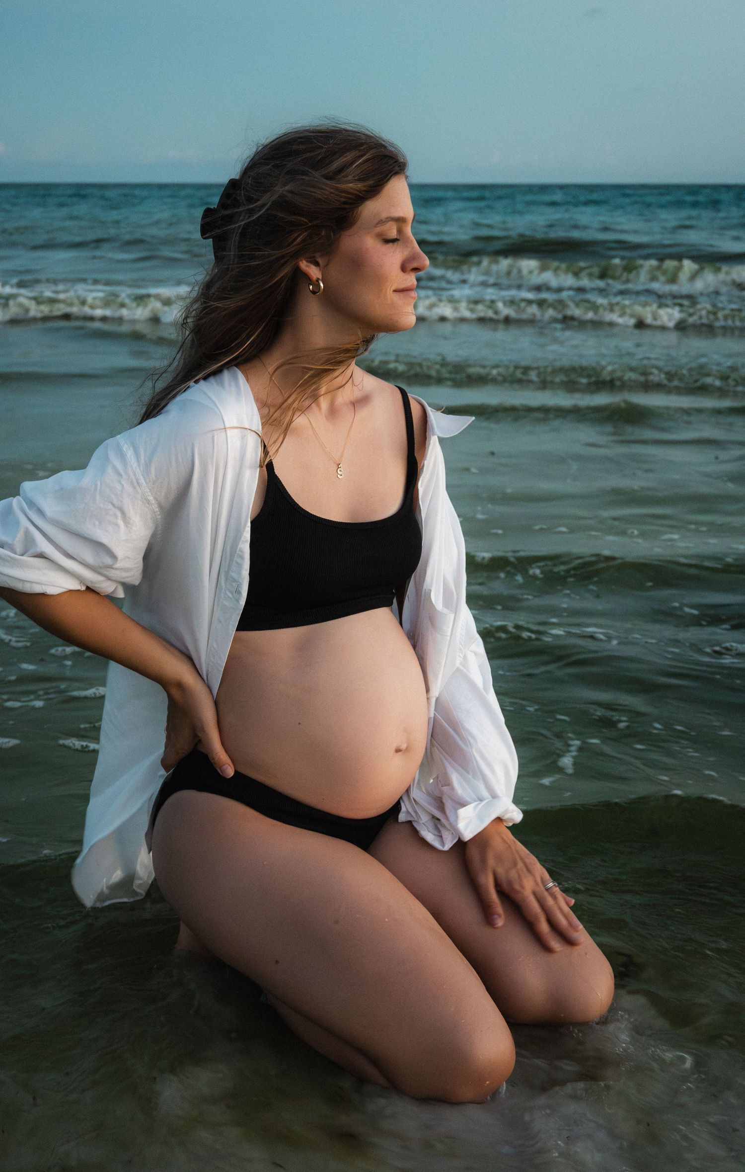 Pregnant woman kneeling in ocean waves wearing black bikini and open white shirt at beach.
