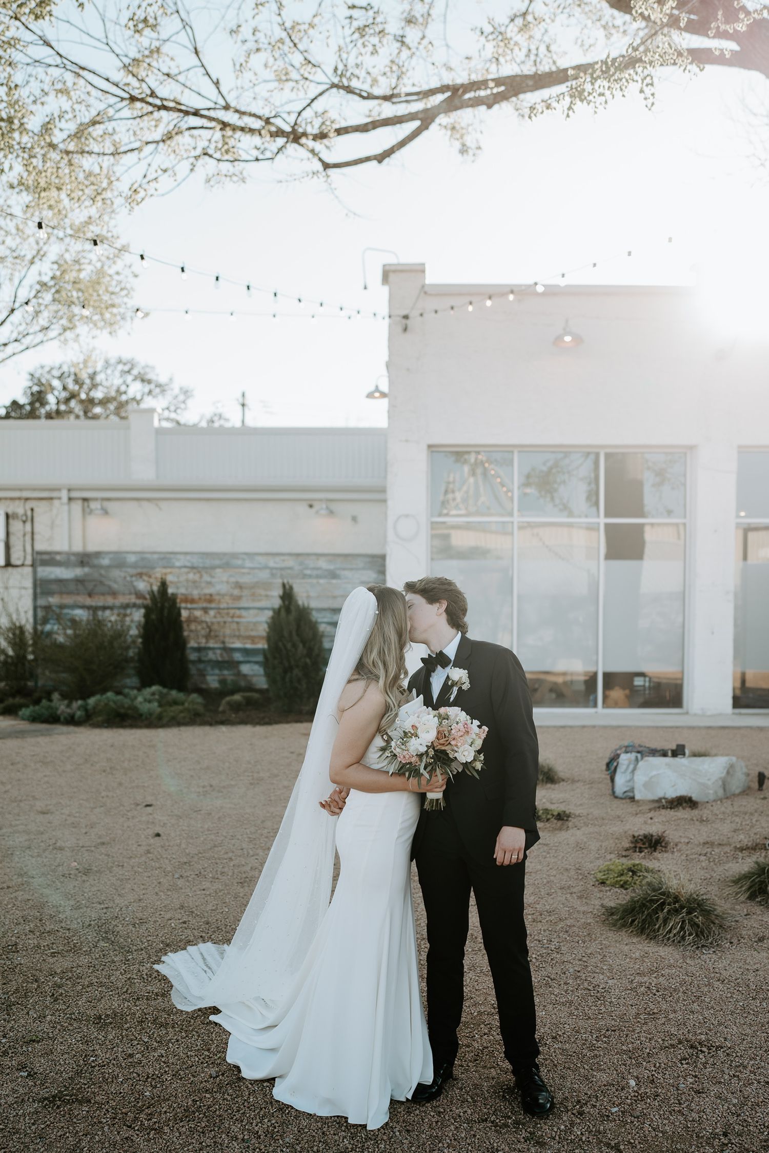 Bride and groom sharing a kiss outdoors with string lights and white building at sunset.