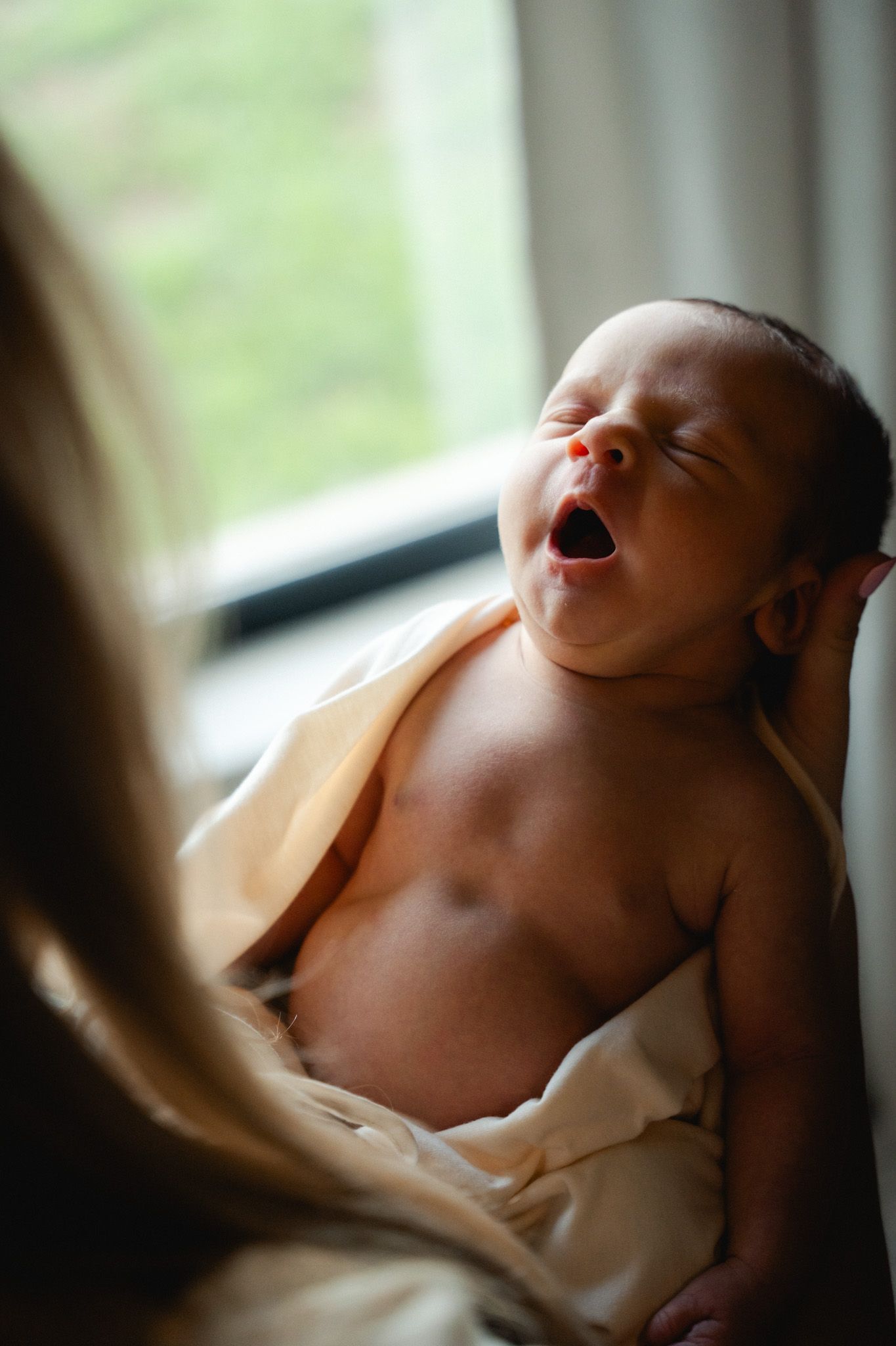 Newborn baby yawning while being held near a bright window, wrapped loosely in a cream blanket.