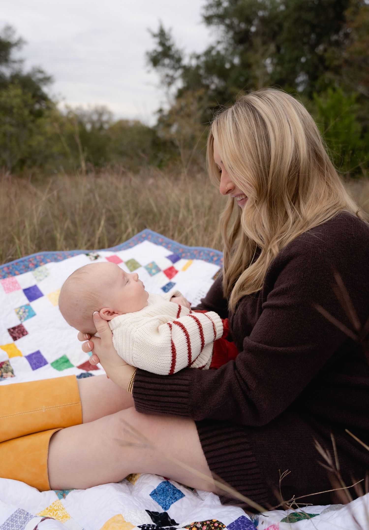 Blonde mother in brown sweater tenderly holding sleeping newborn baby on colorful patchwork quilt outdoors.