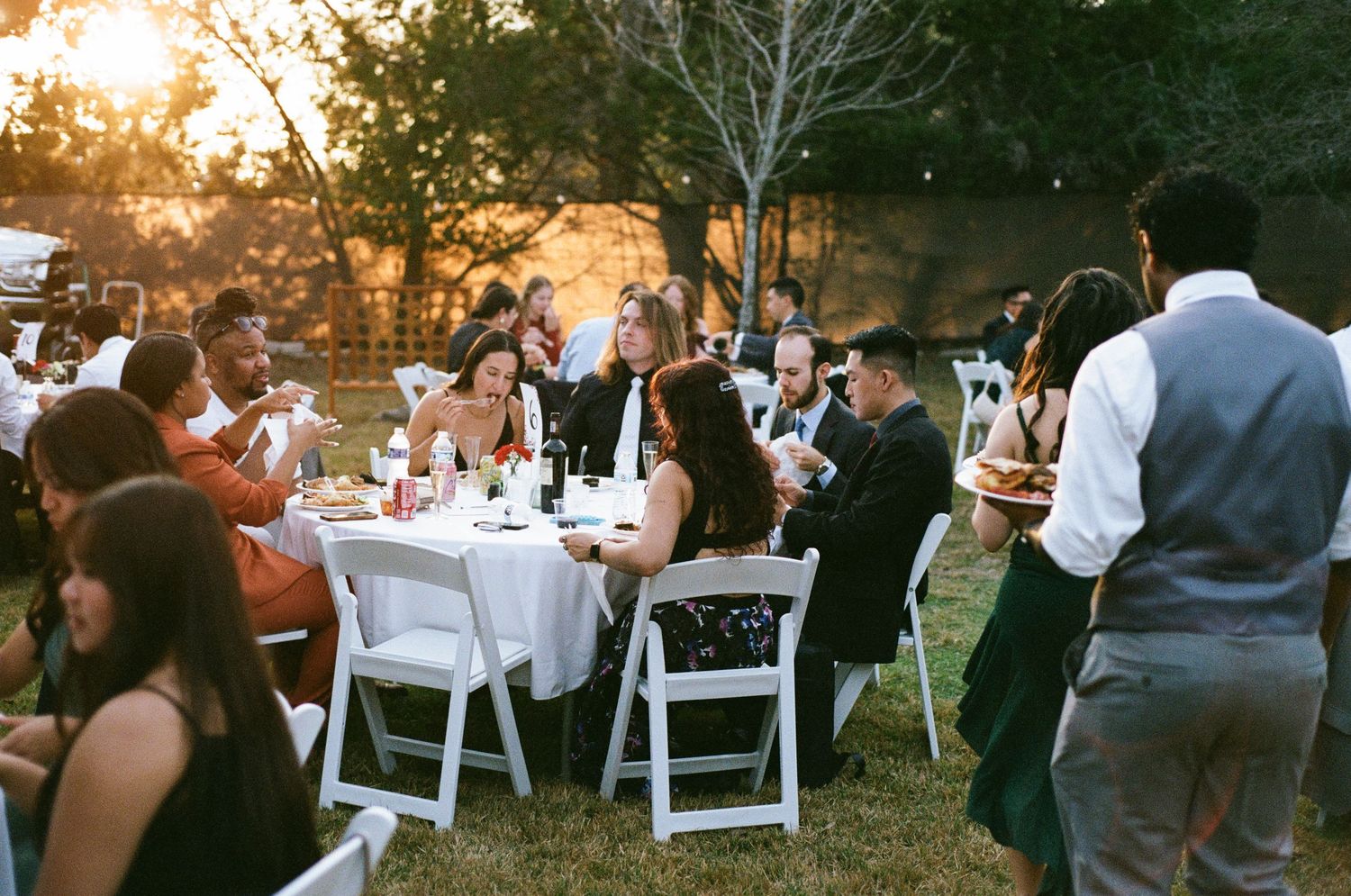 Wedding guests dine at white round tables on a lawn at golden hour.