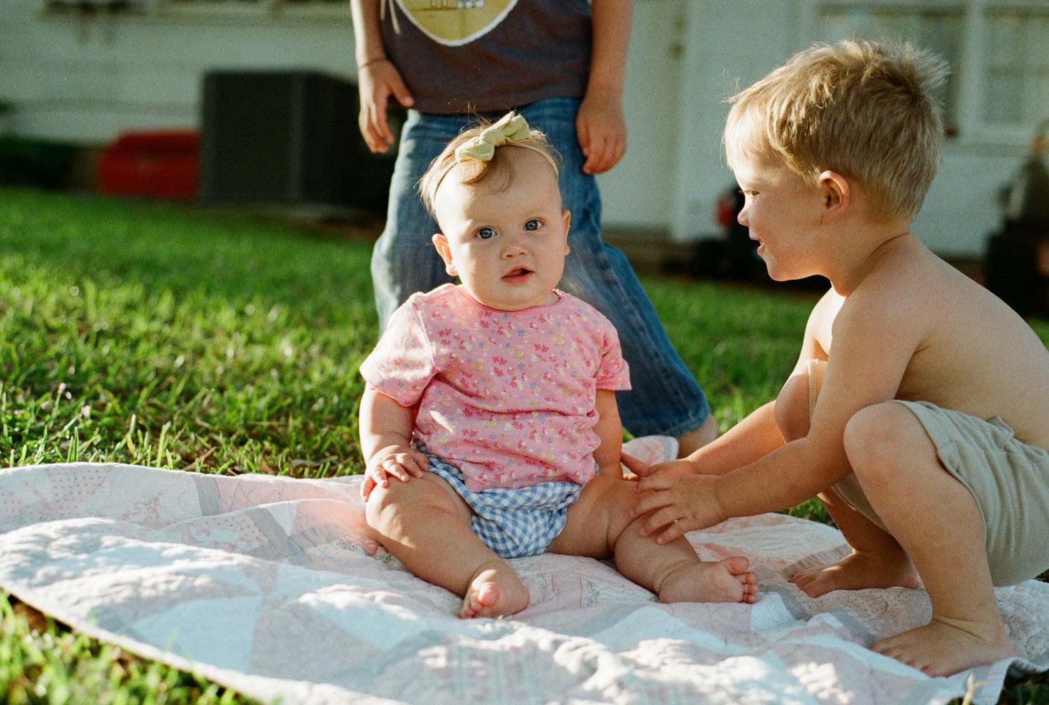 Two young siblings sit on a quilt outdoors as an older boy interacts with a baby girl in a pink outfit.