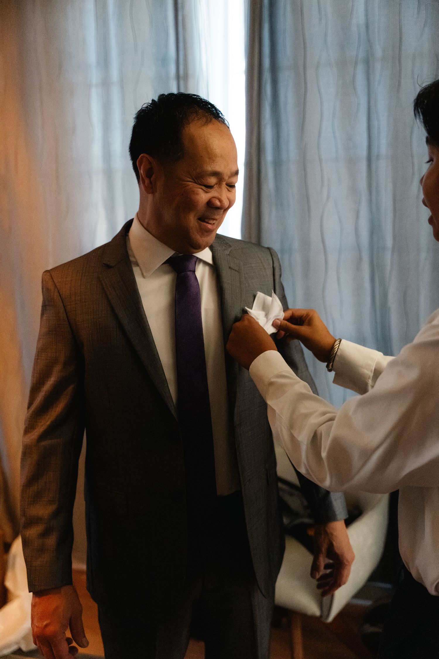 Smiling groom gets boutonniere pinned to gray suit jacket before wedding ceremony.