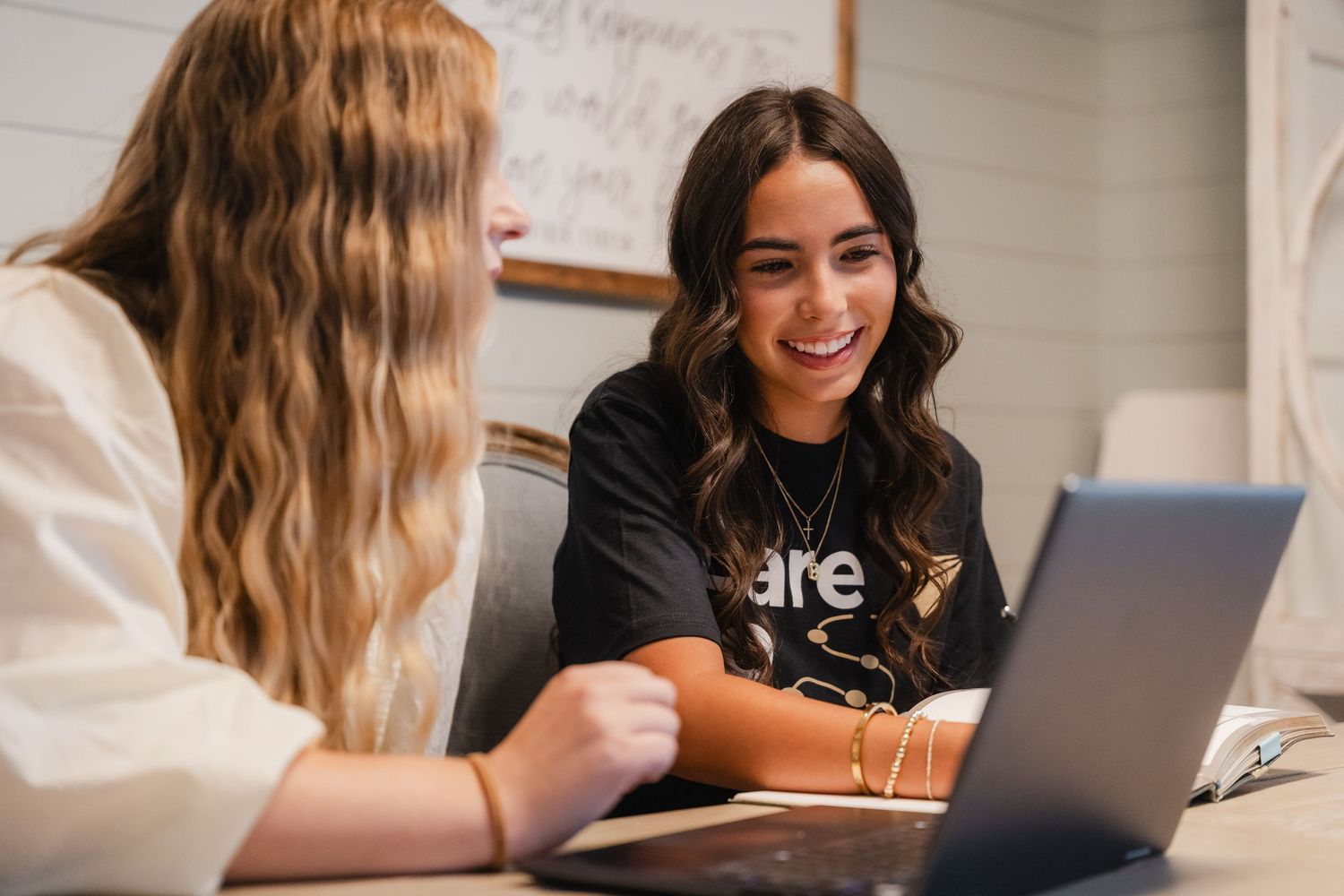 Two young women smiling and collaborating on a laptop at a wooden table indoors.