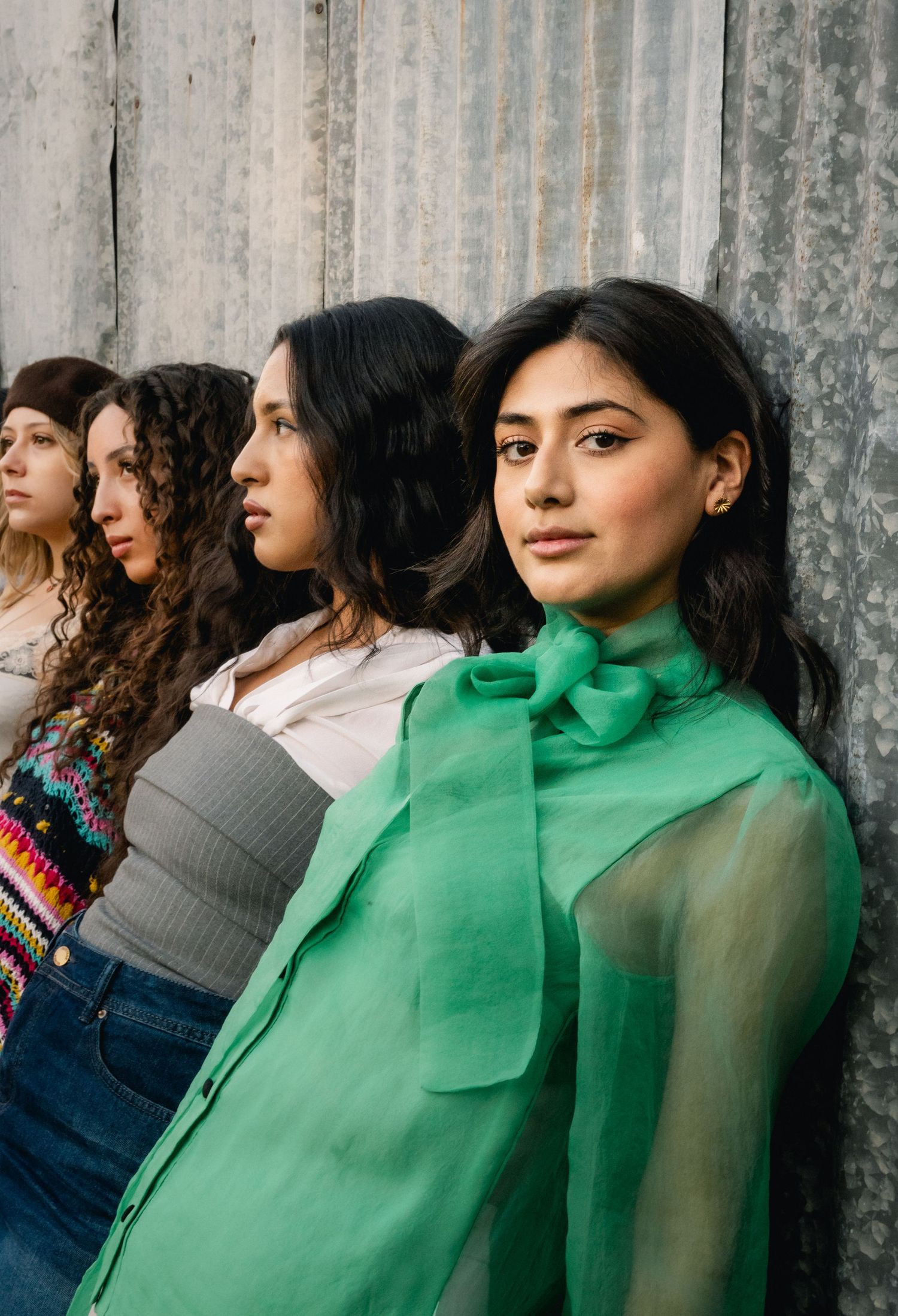 Four young women standing against a corrugated metal wall, front woman wearing a green blouse.
