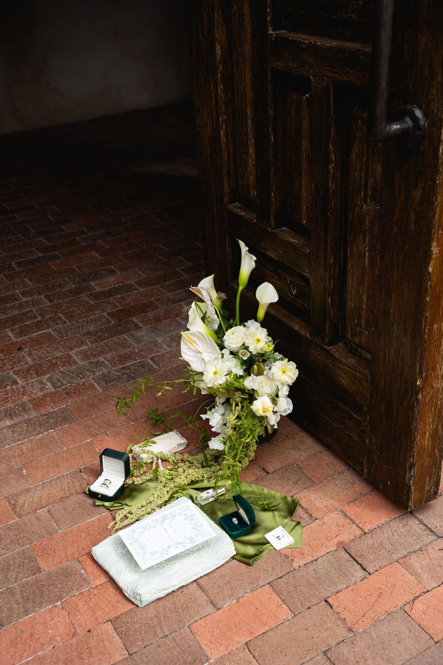 Wedding bouquet of white calla lilies and roses with rings and vow books on brick floor by wooden door.