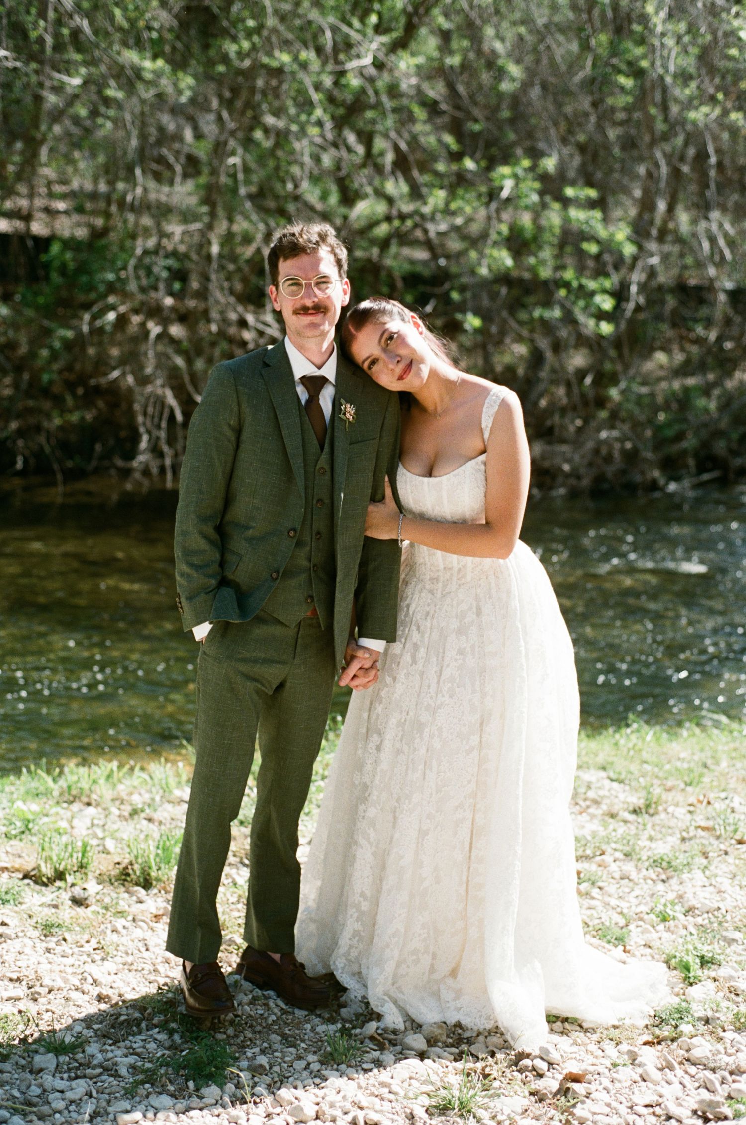Bride and groom holding hands and smiling by a creek in a lush green outdoor wedding setting.