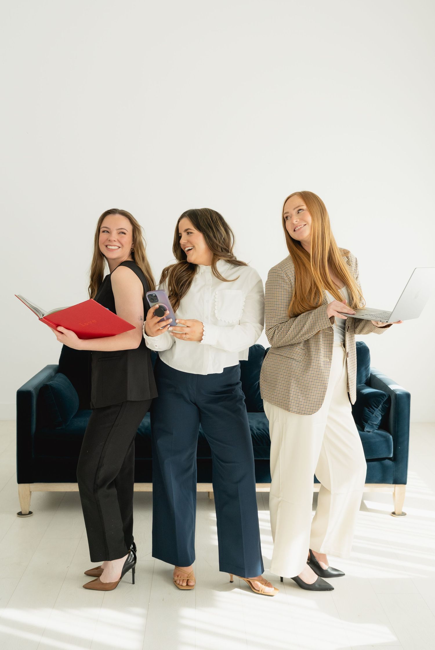 Three smiling professional women standing with laptop, phone, and binder in a bright white studio.