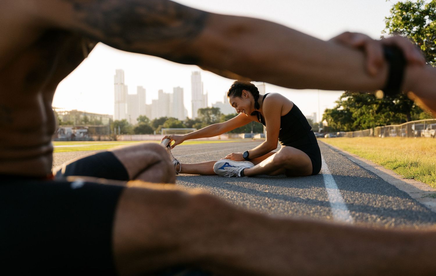 Two athletes stretching on an outdoor running track with a city skyline in the background.