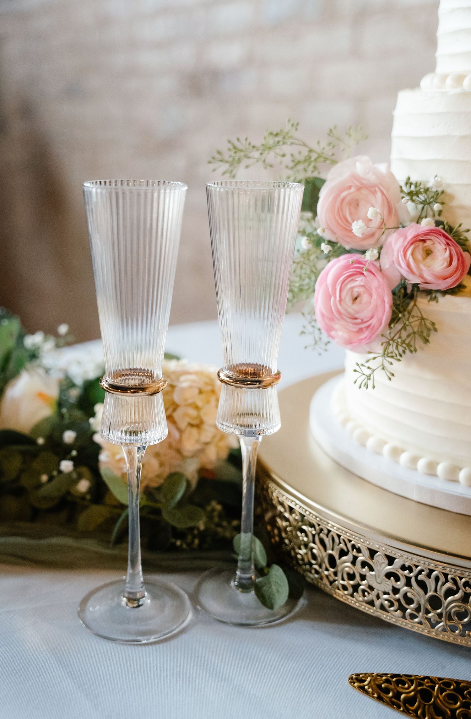 Two elegant ribbed champagne flutes with gold rings beside a white wedding cake adorned with pink ranunculus flowers.