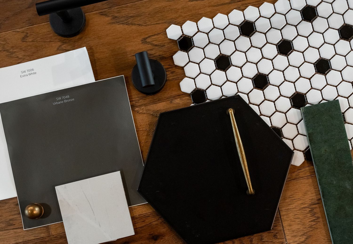 Flat lay of interior design mood board with black hexagon tile, white mosaic tile, dark gray paint swatch, and brass pen on wood.