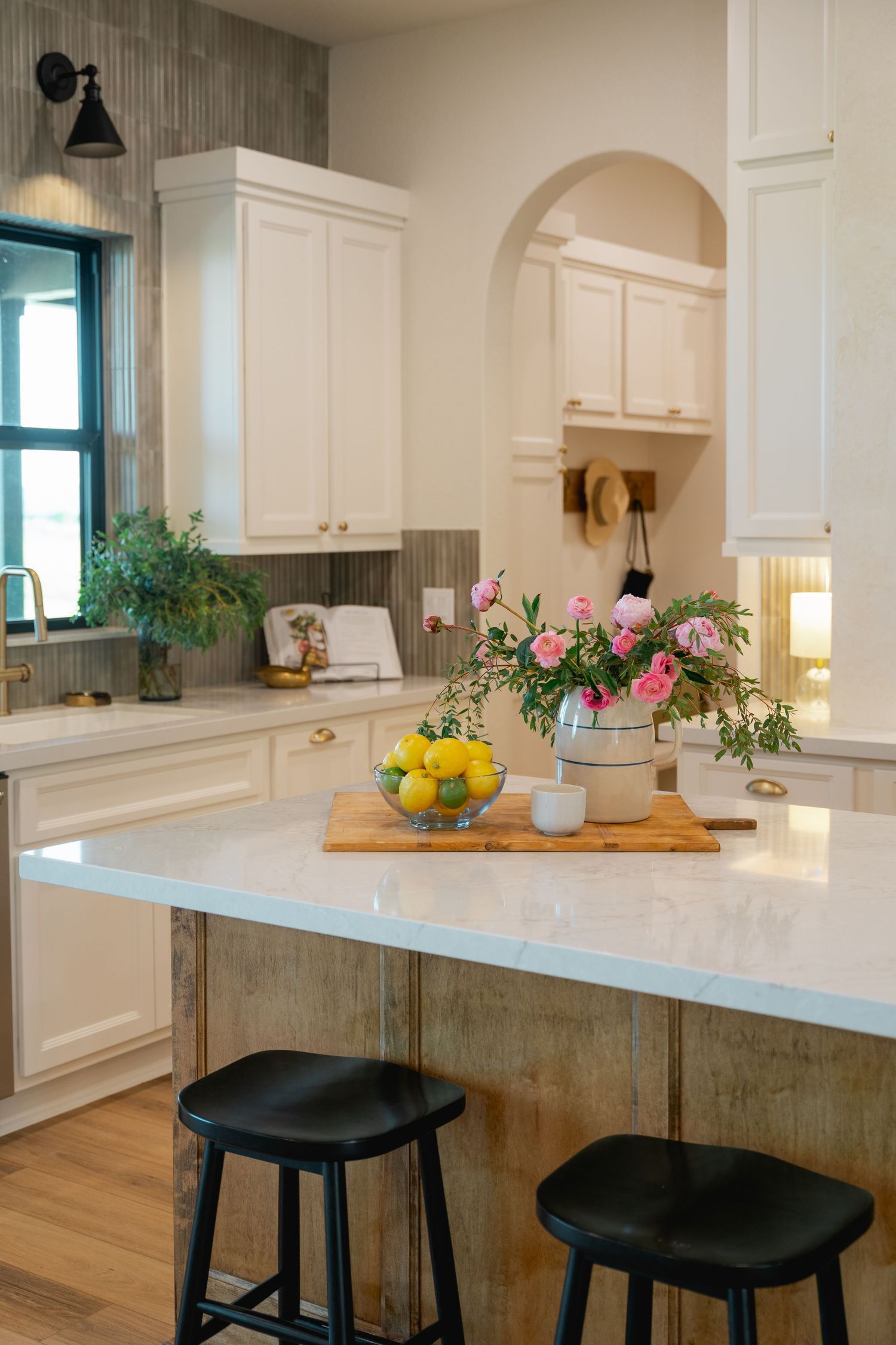 White kitchen with marble island, black stools, pink floral arrangement, and lemons on cutting board.