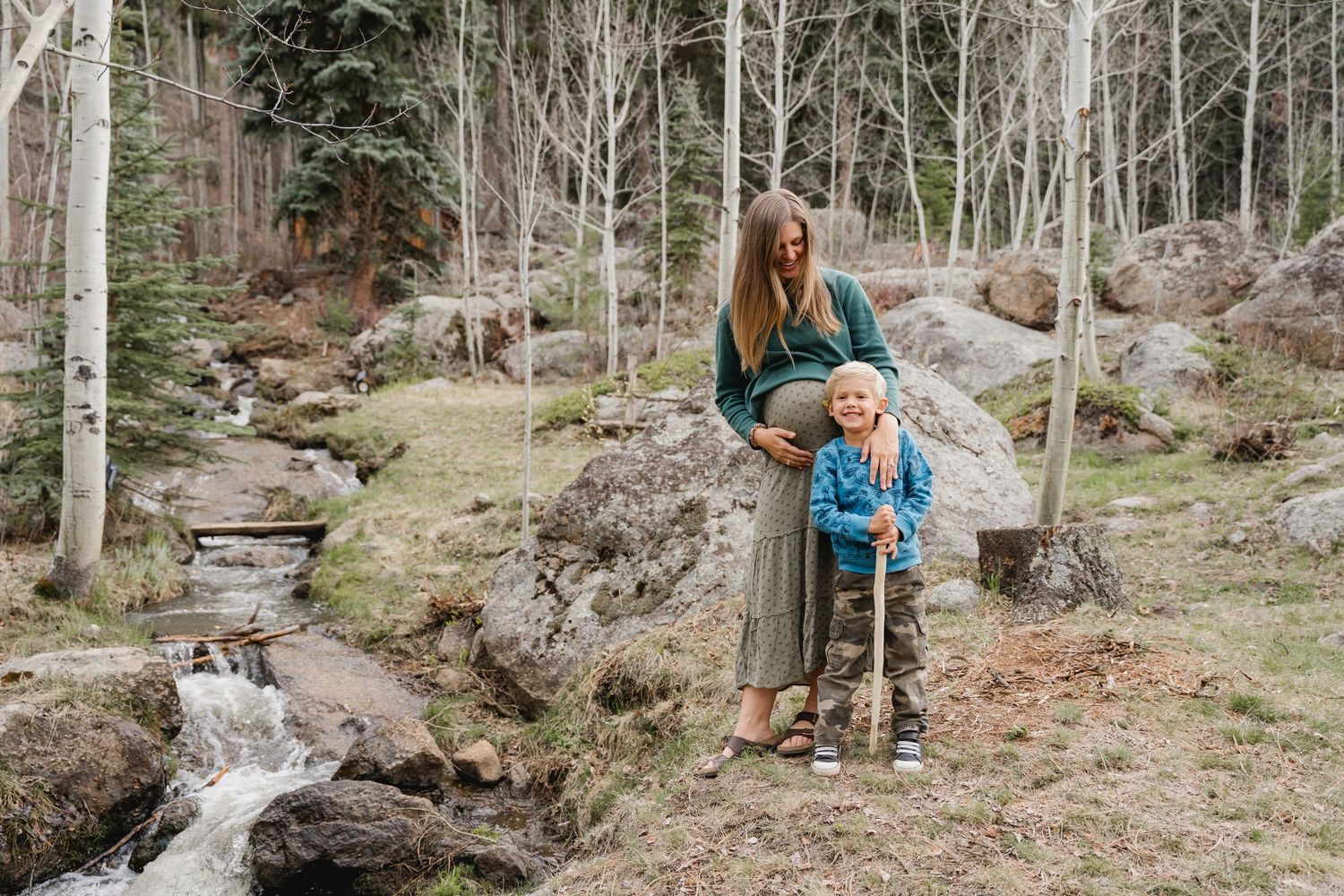 Pregnant woman in green sweater and skirt poses with toddler by rocky stream in aspen forest.