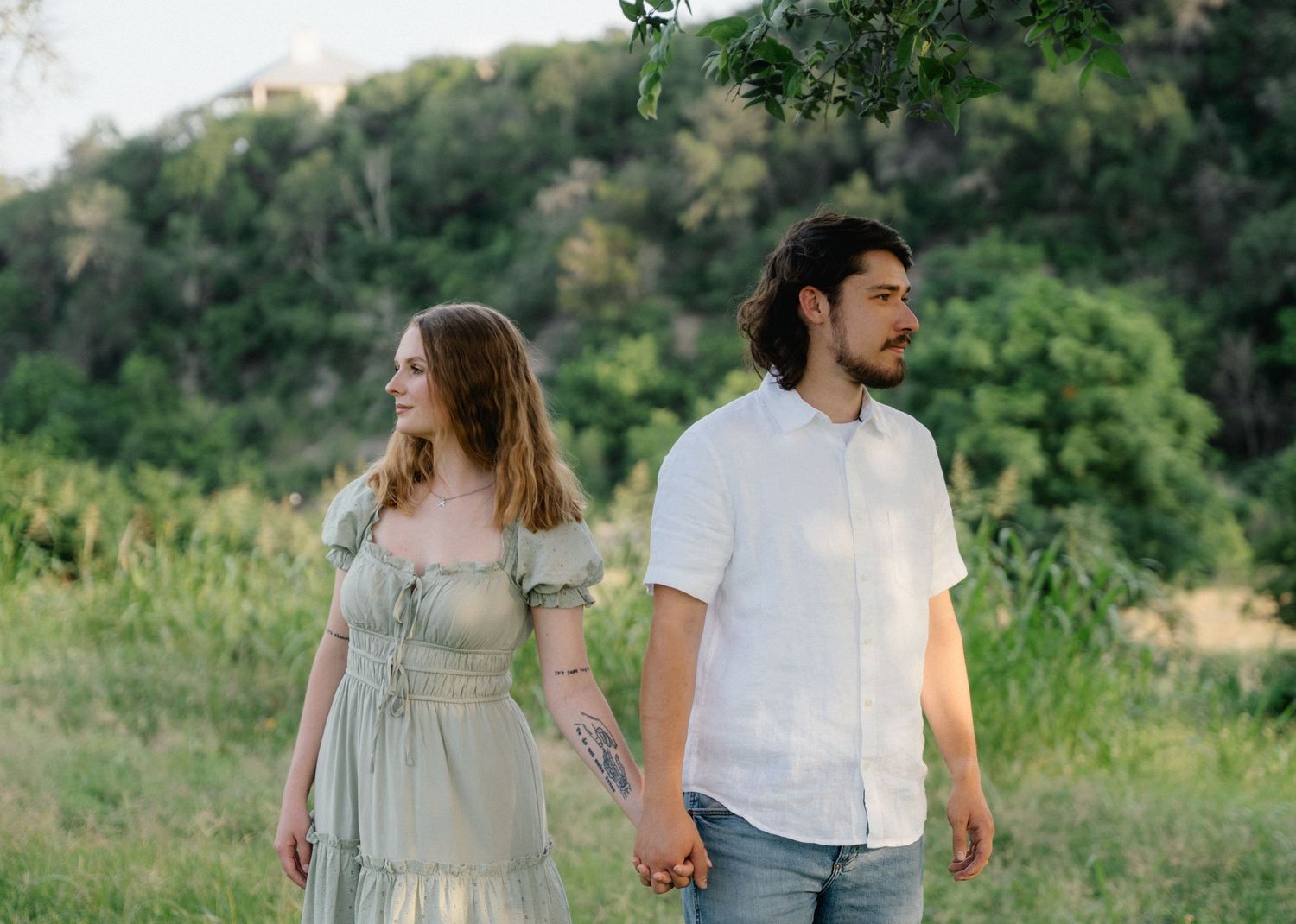 A young couple holds hands while walking through a lush green meadow, dressed in elegant casual attire.