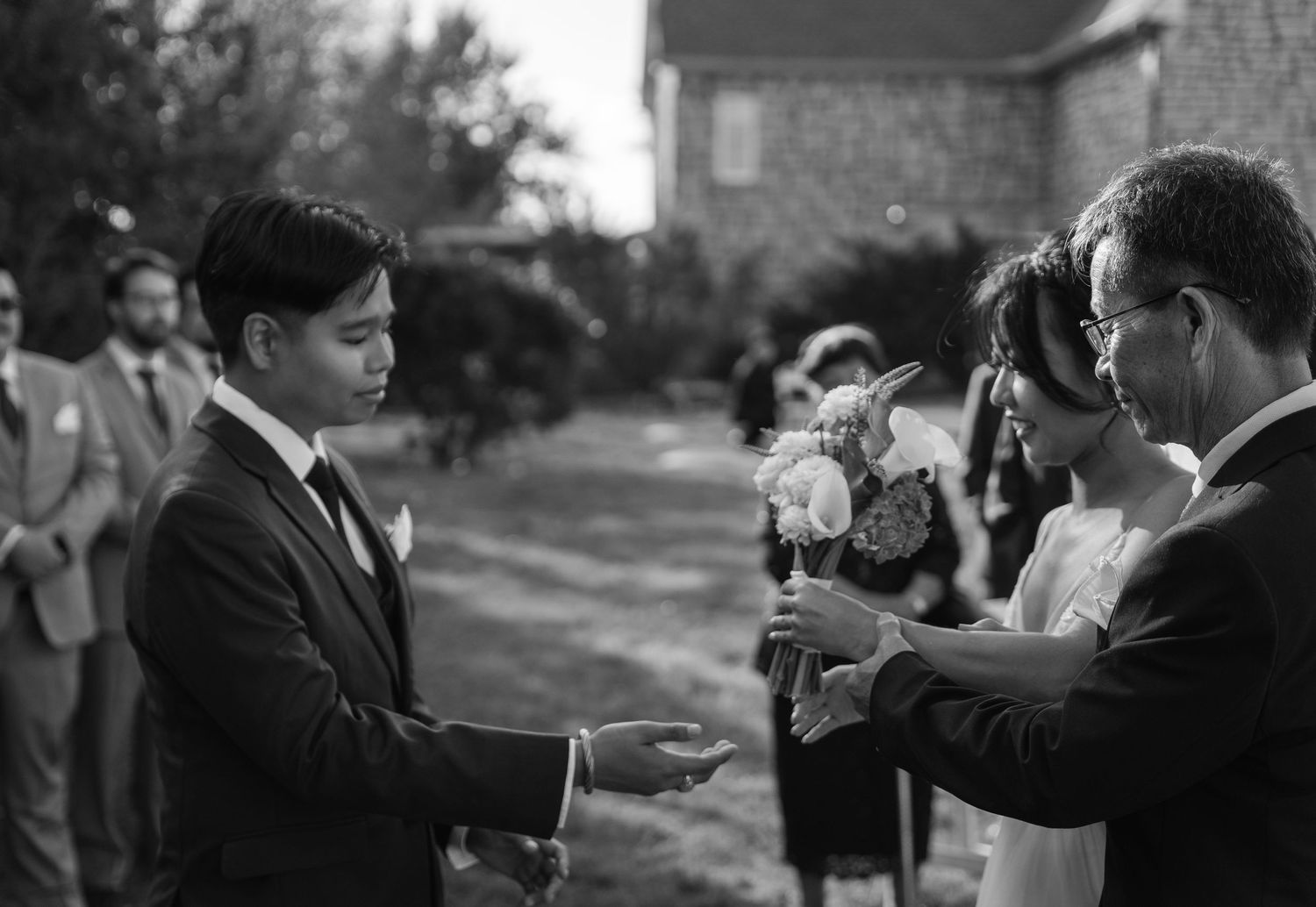 Black and white photo of groom receiving bride from father at outdoor wedding ceremony.