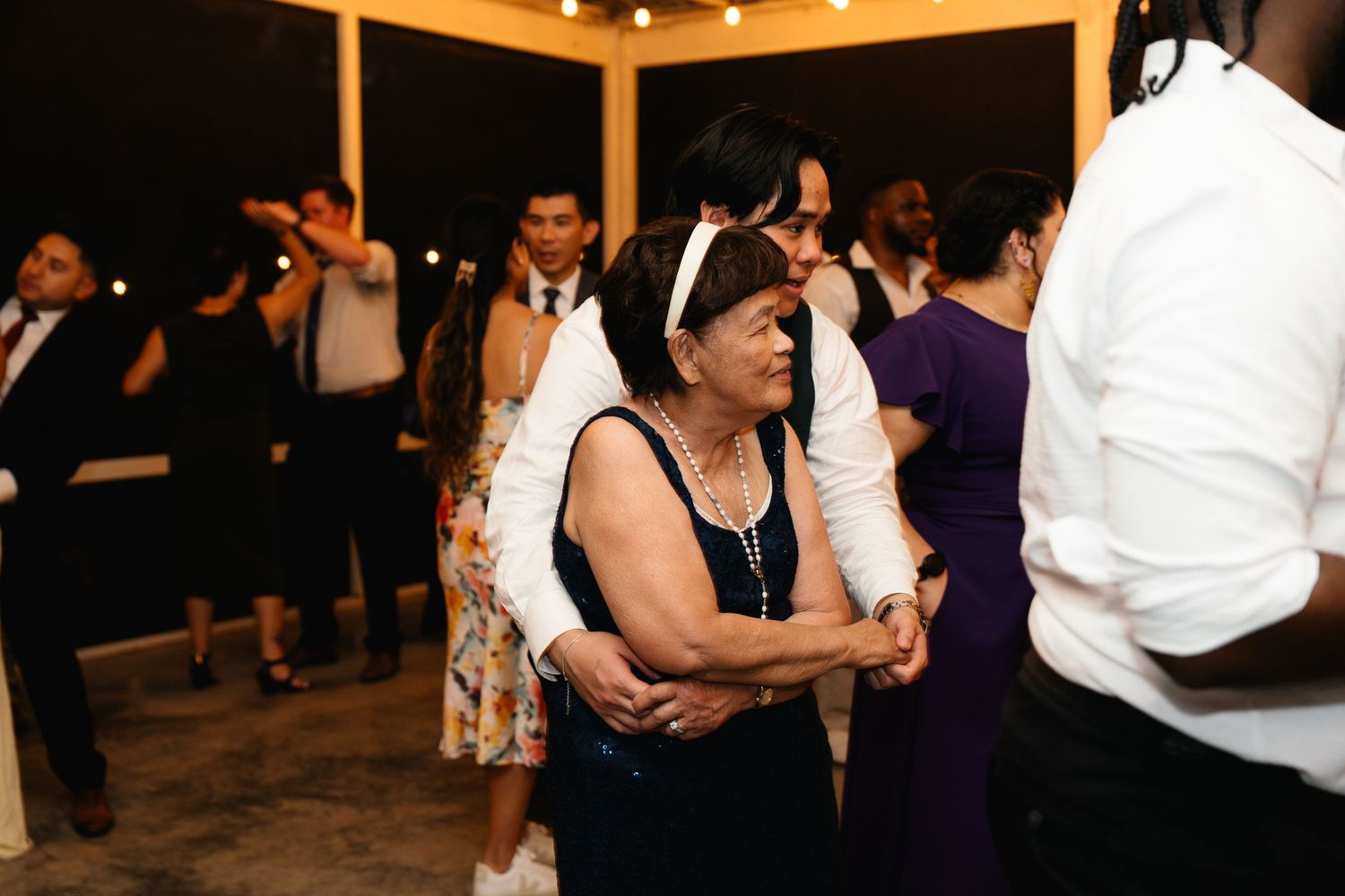Elderly woman dances enthusiastically among wedding guests at lively outdoor evening reception.