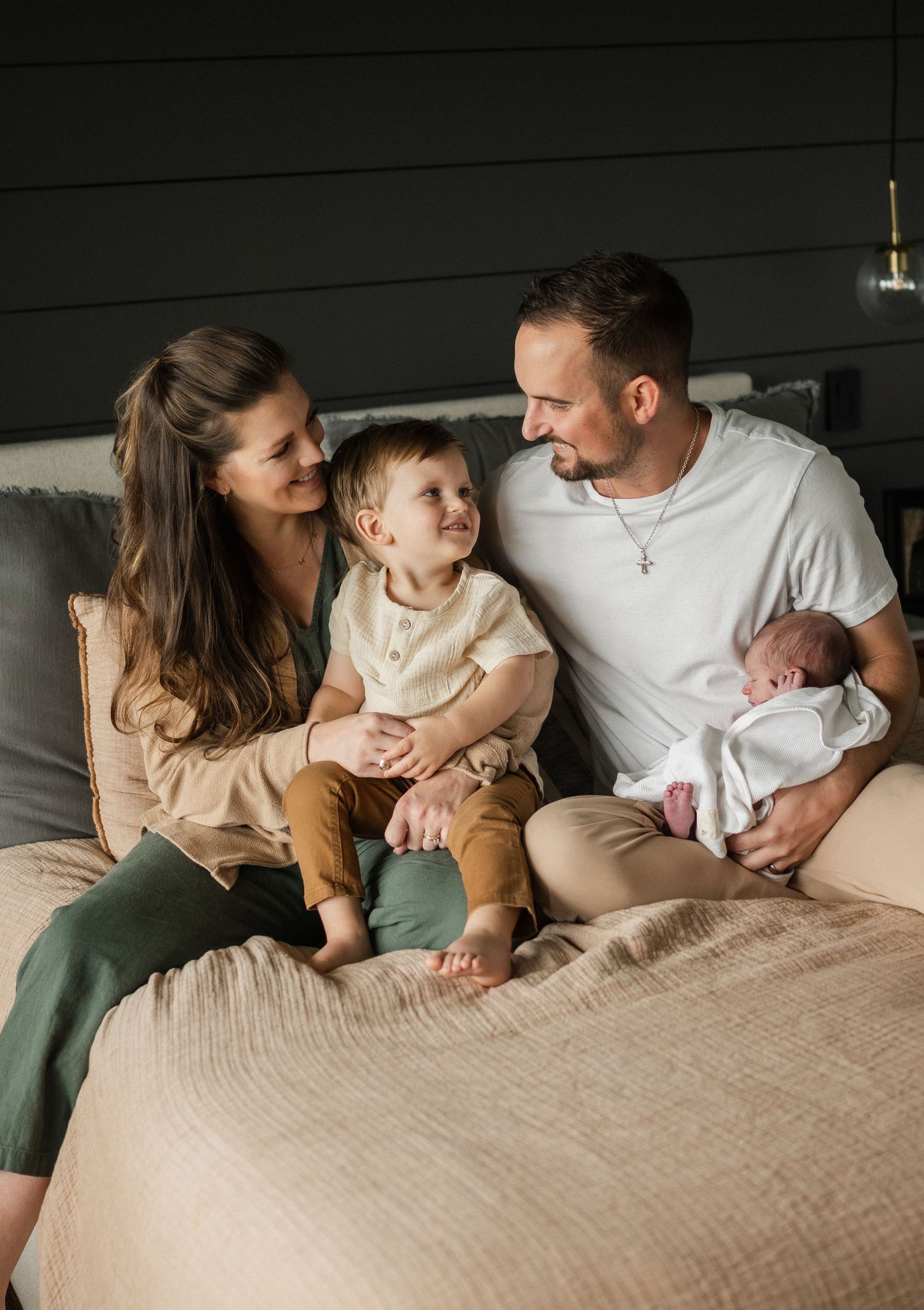 Happy family of four with toddler and newborn sitting together on bed in dark bedroom.