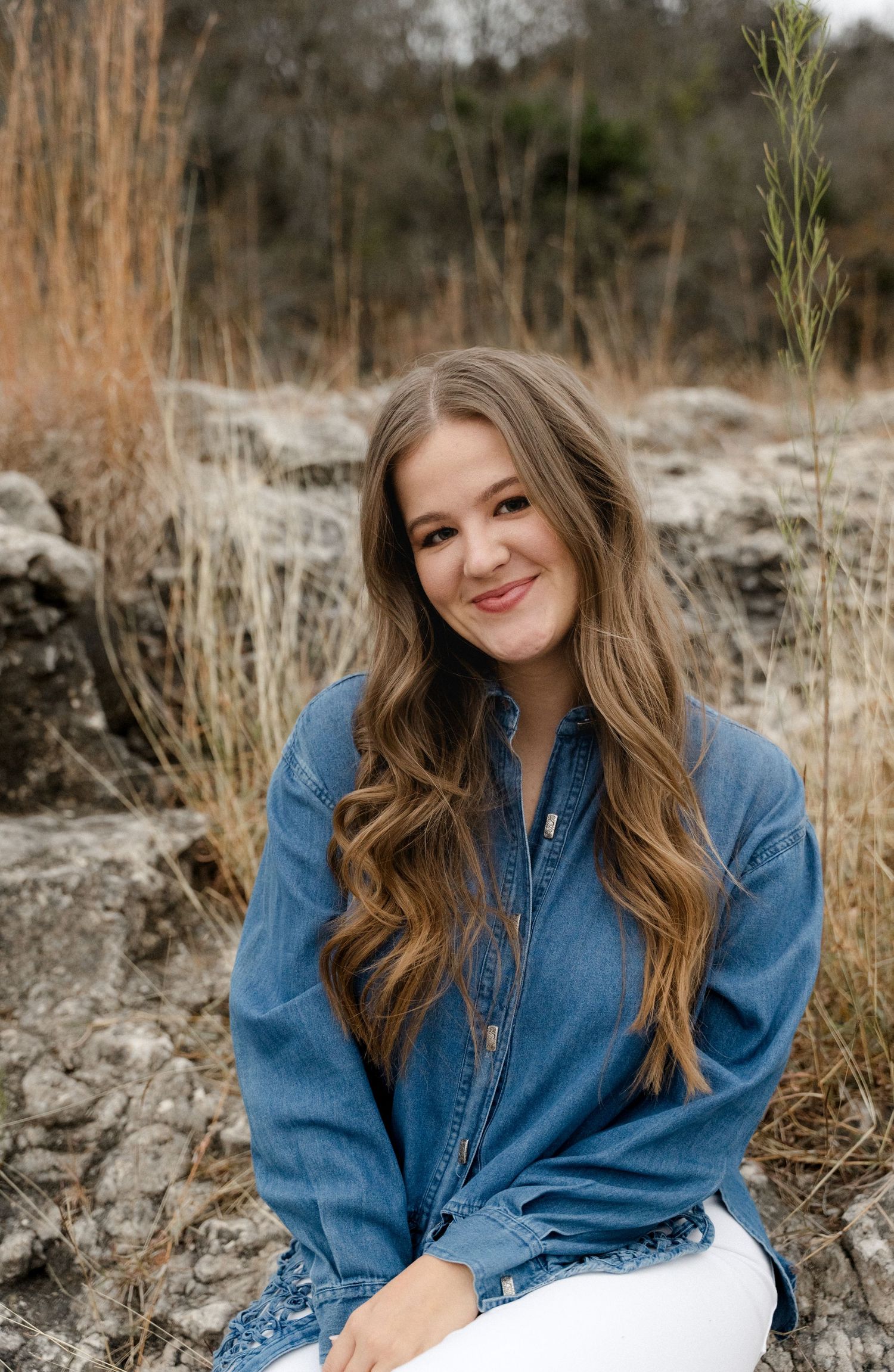 Smiling young woman in a denim shirt sitting on rocks in a natural outdoor setting.