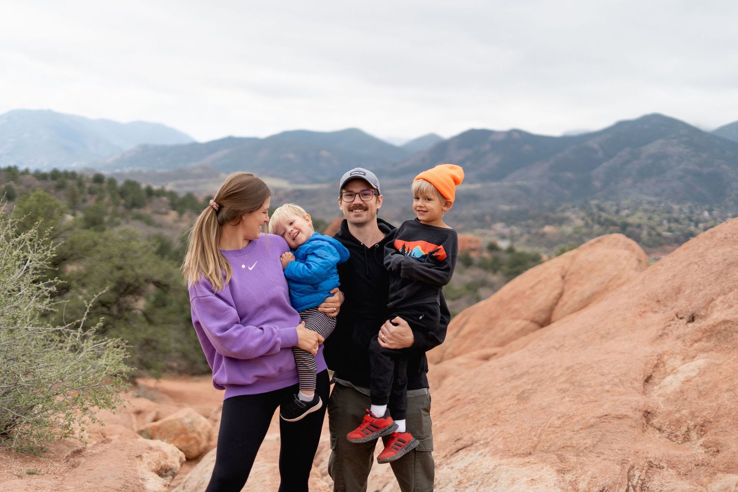 Happy family with two toddlers posing at Garden of the Gods with red rocks and mountains backdrop.