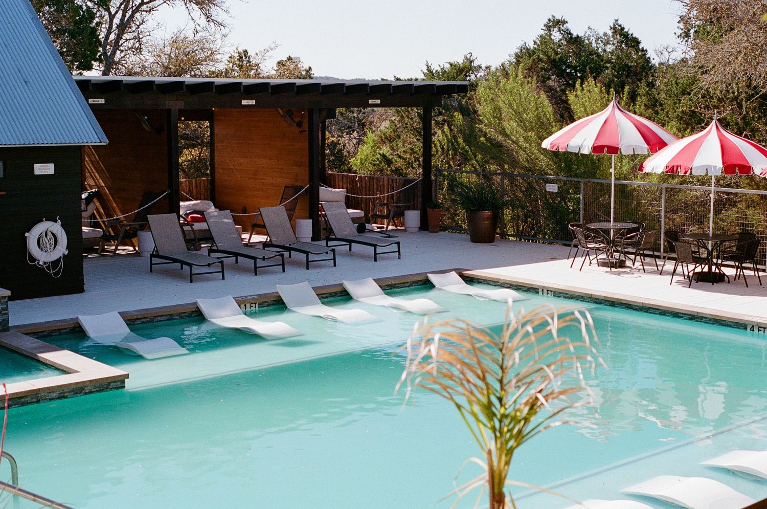 Outdoor resort pool with lounge chairs, red striped umbrellas, and lush greenery on a sunny day.