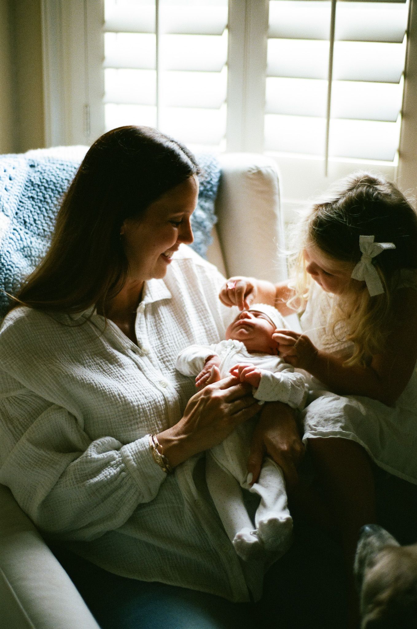 A mother sitting in a white chair holds her newborn while a young blonde girl in white dress looks on lovingly.