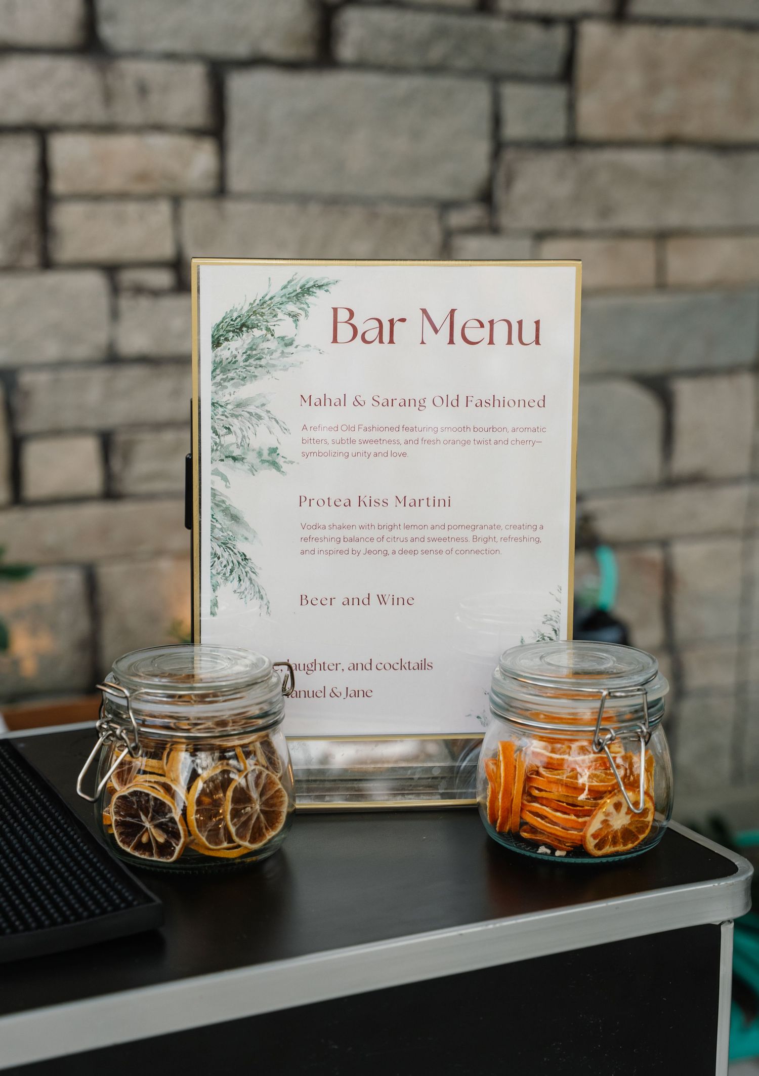 Elegant wedding bar menu display with signature cocktails flanked by dried orange slice jars.