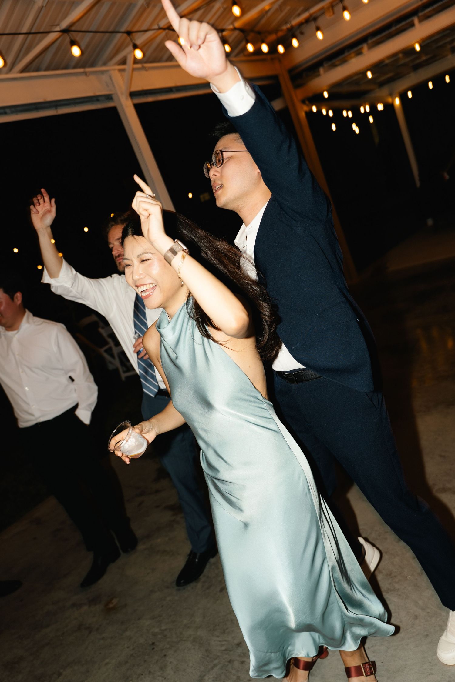 Wedding guests dance joyfully under string lights, woman in sage green dress twirling.