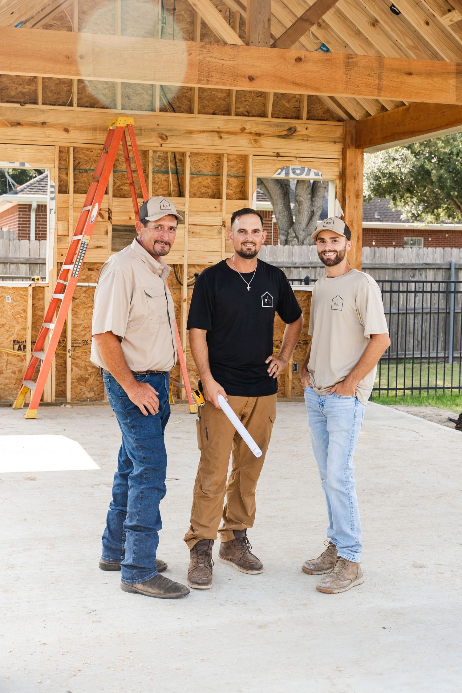 Three construction workers smiling at a building site with lumber framing and an orange ladder in the background.