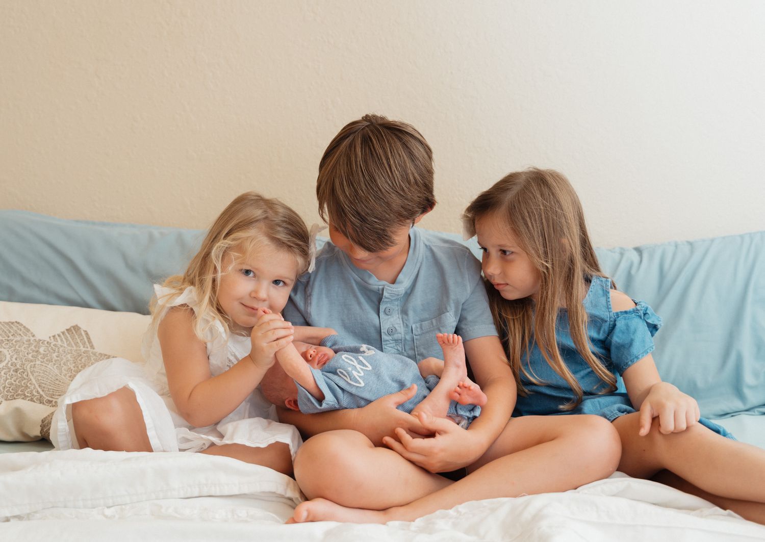 Three siblings sit on a bed, curiously examining their newborn baby sibling together.