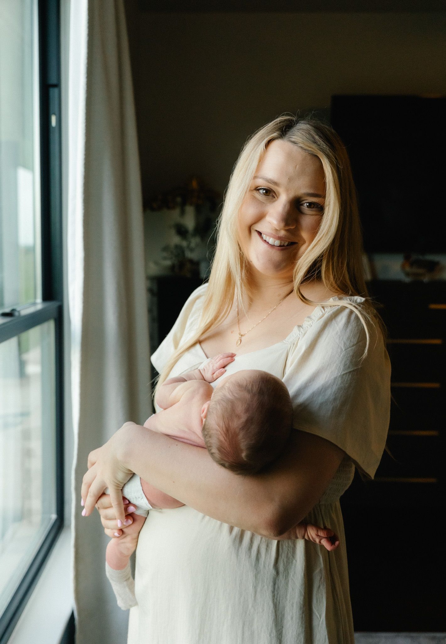 Smiling blonde mother in cream dress holding sleeping newborn baby while standing near a window.