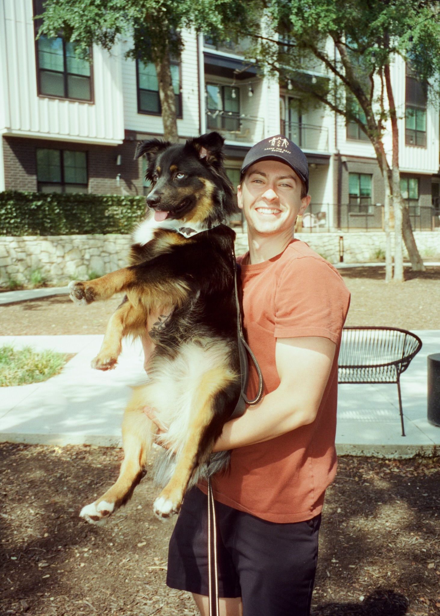Smiling man holding a large tri-color Collie dog outdoors near an apartment building.