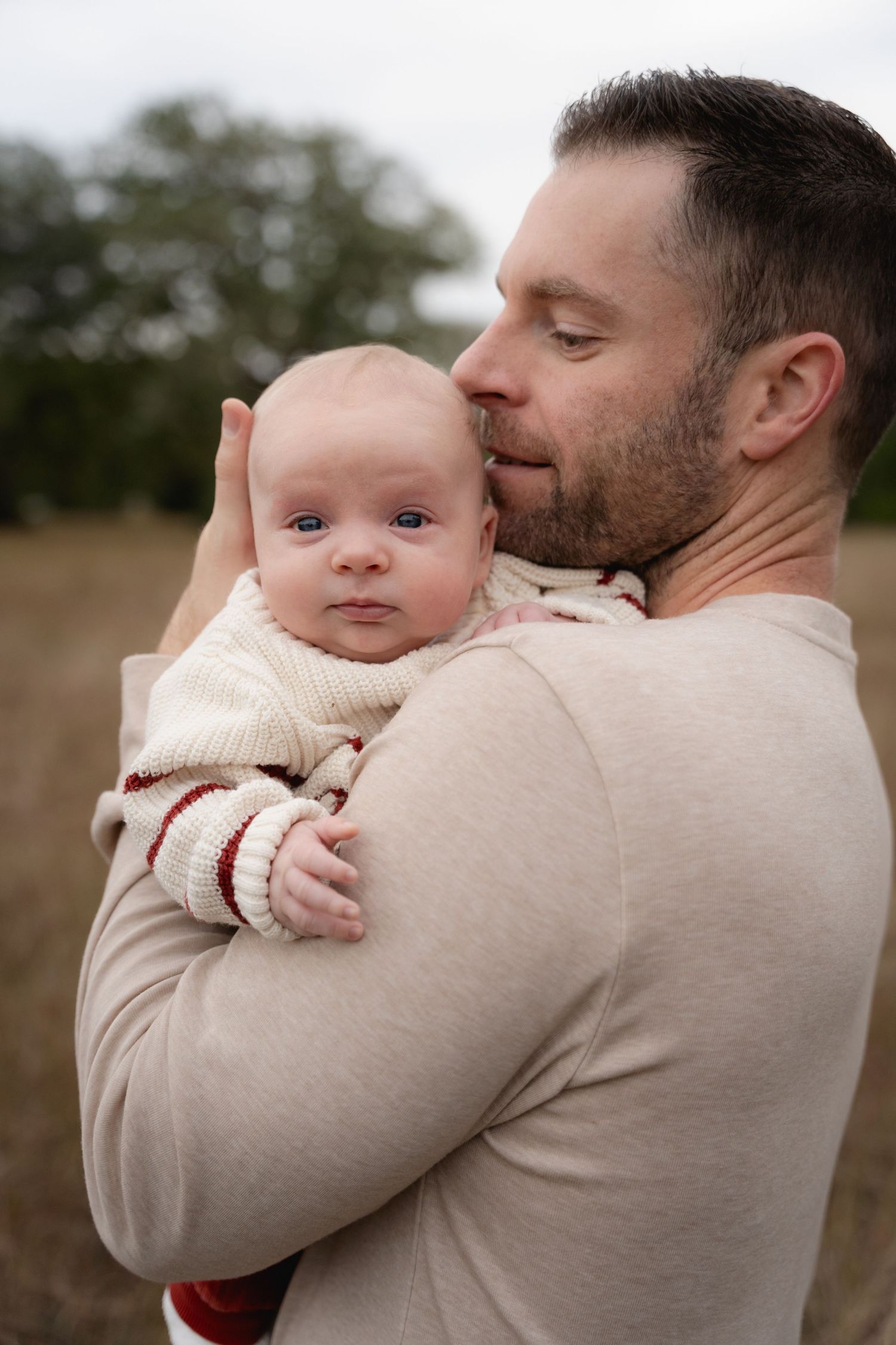 Loving father in beige shirt holding alert newborn baby close to his cheek in autumn field.