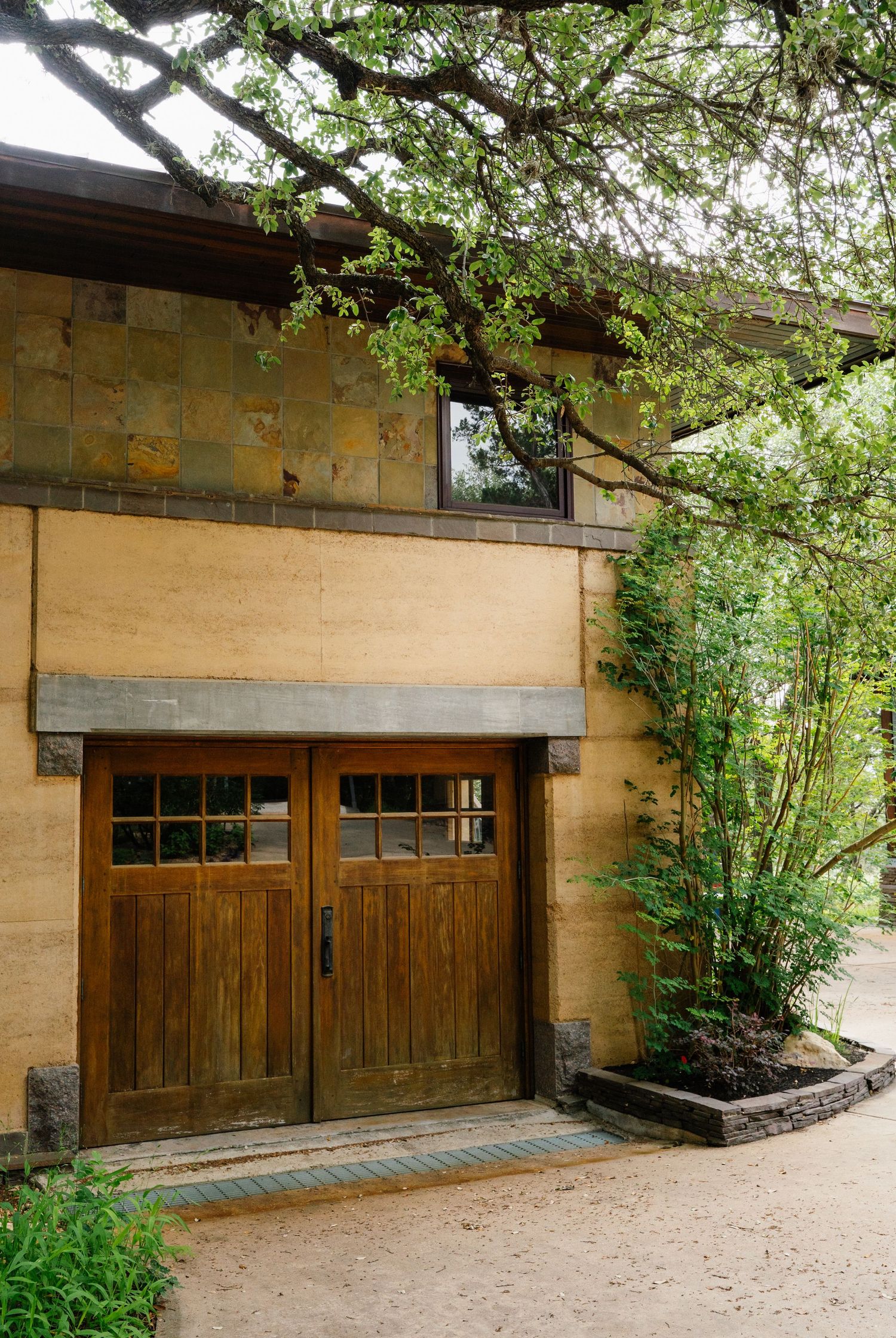 Rustic wooden double doors with glass panels on a rammed earth building surrounded by lush green landscaping.