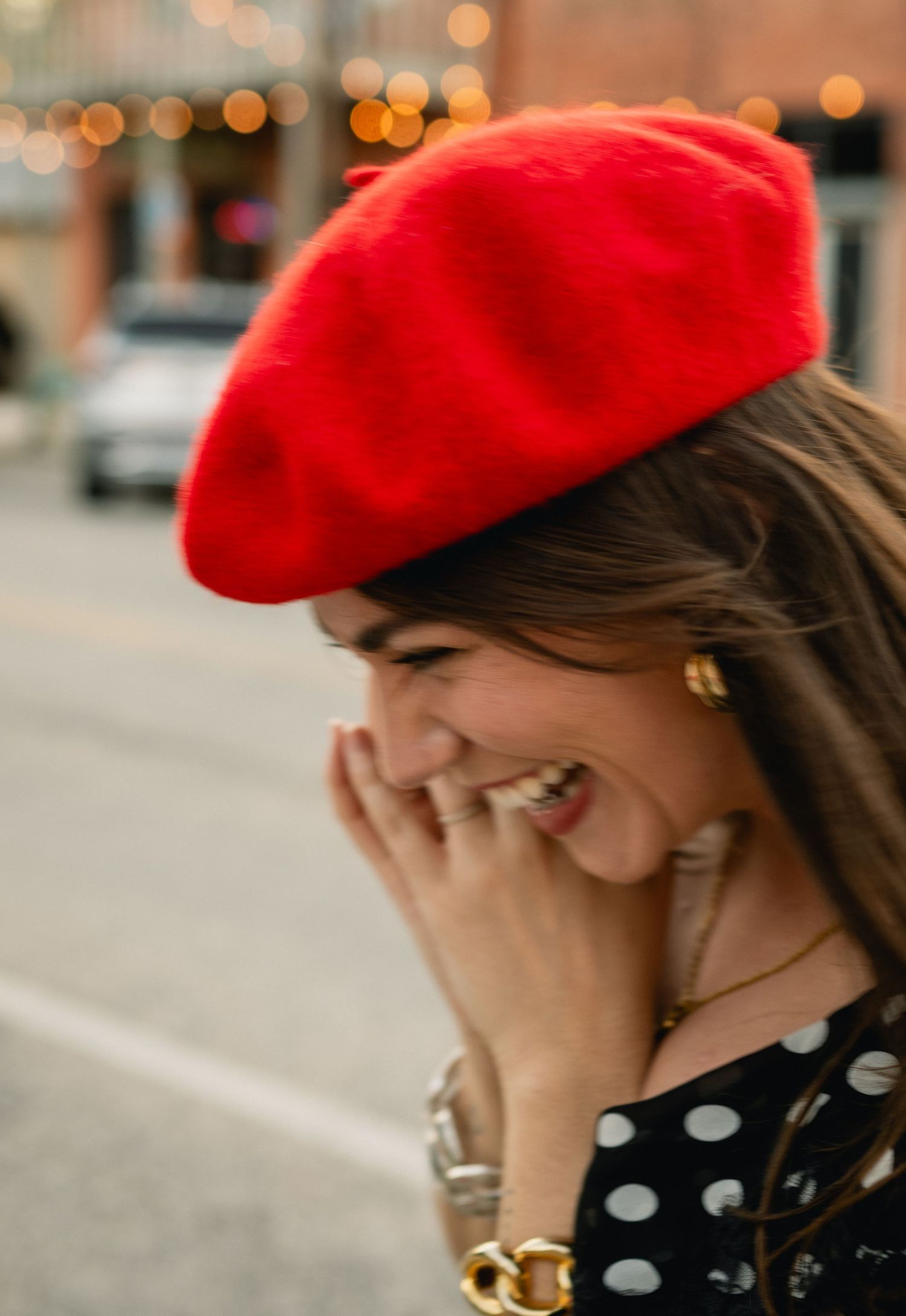 Laughing woman wearing a bright red beret and polka dot dress with gold jewelry outdoors.