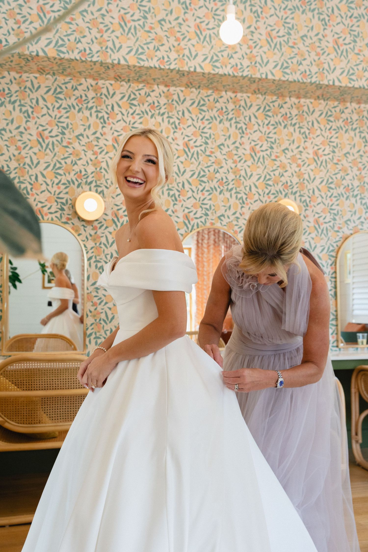 Smiling blonde bride in off-shoulder white ball gown being buttoned up in floral wallpapered dressing room.