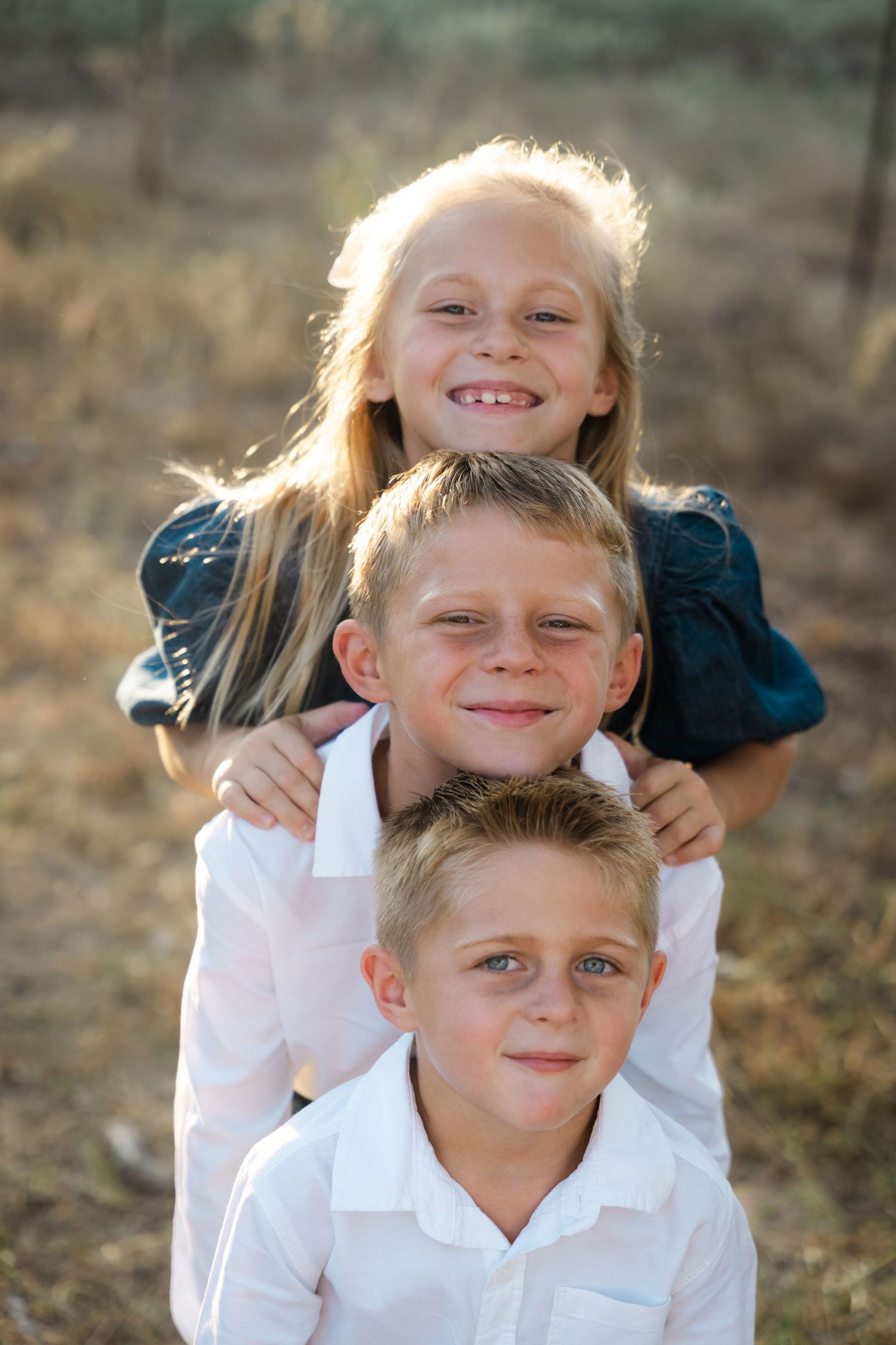Three blonde siblings pose together smiling in a sun-drenched outdoor setting.