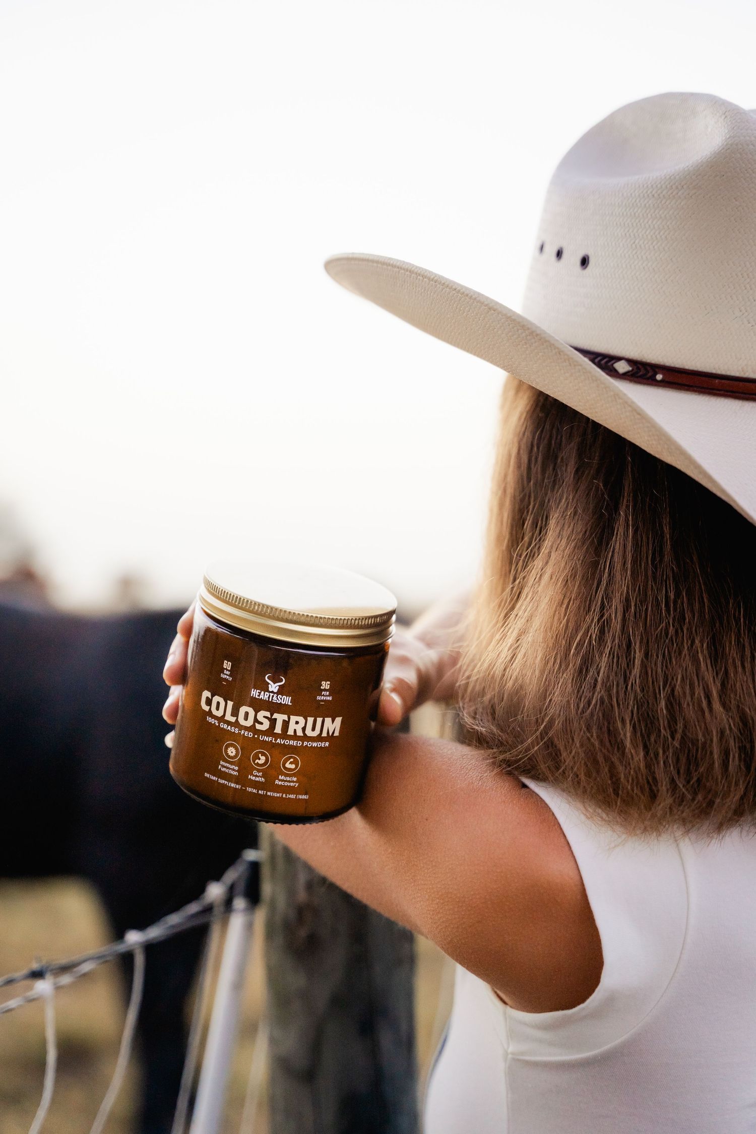 Woman in white cowboy hat holding a jar of Colostrum supplement near a fence.
