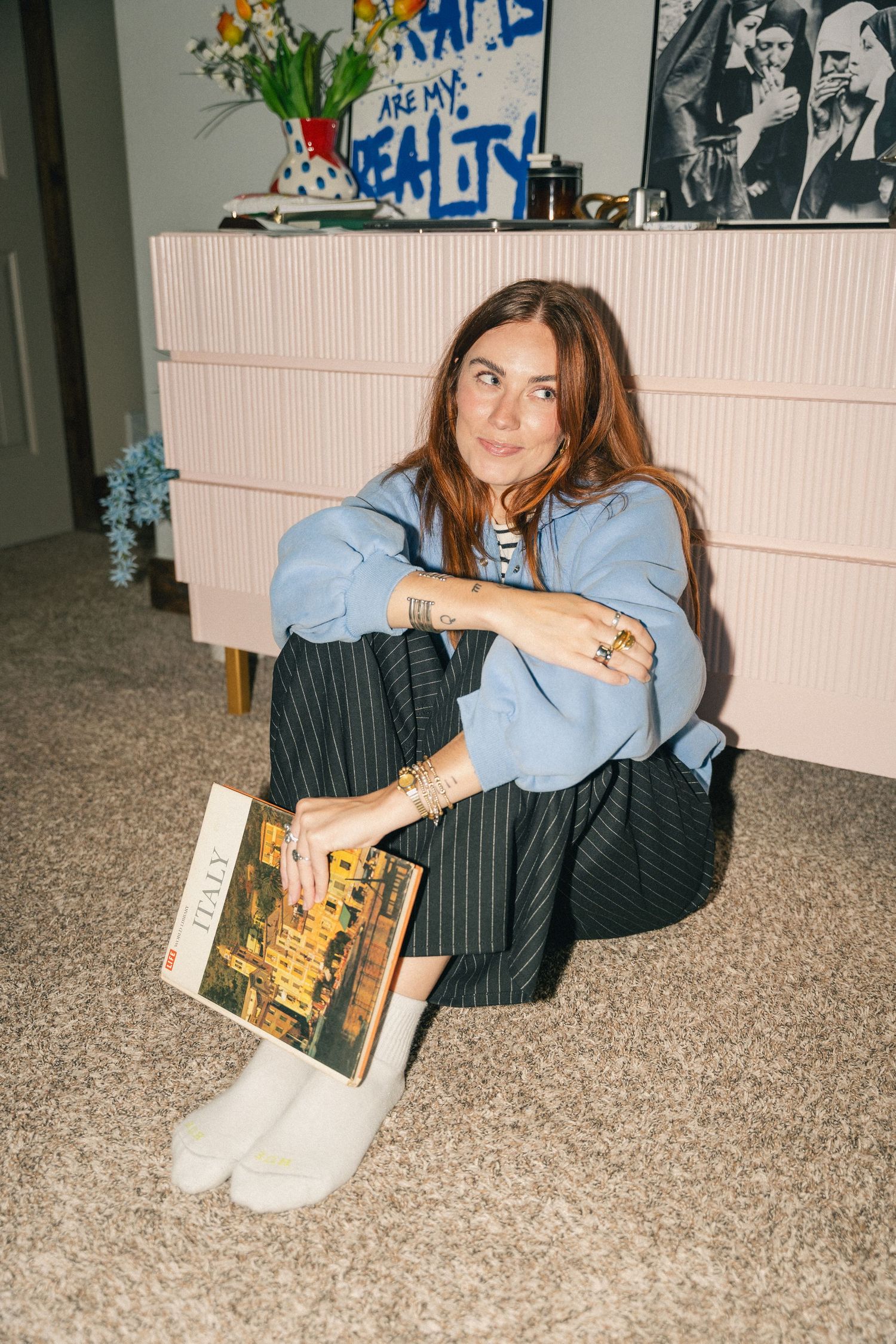 Young woman sitting on floor holding a book, wearing blue shirt and pinstripe pants, leaning against a pink dresser.