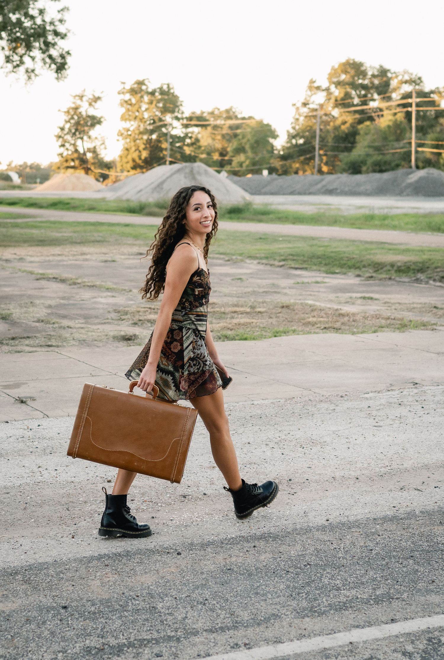 Young woman in floral dress and boots carrying a brown vintage suitcase across a parking lot.