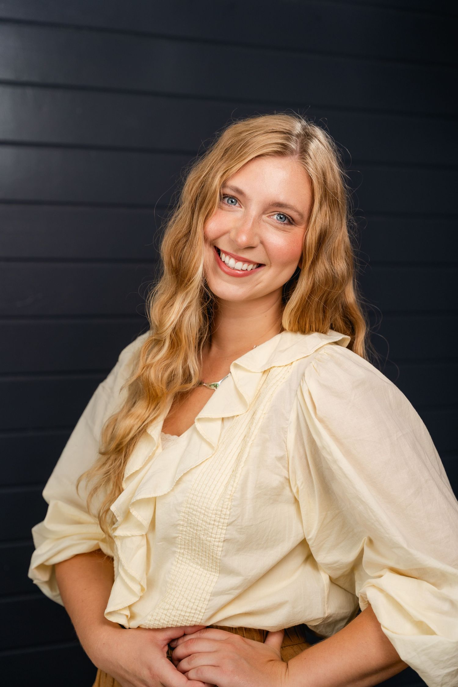 Smiling young woman with blonde wavy hair wearing a cream ruffled blouse, posing against a dark wall.