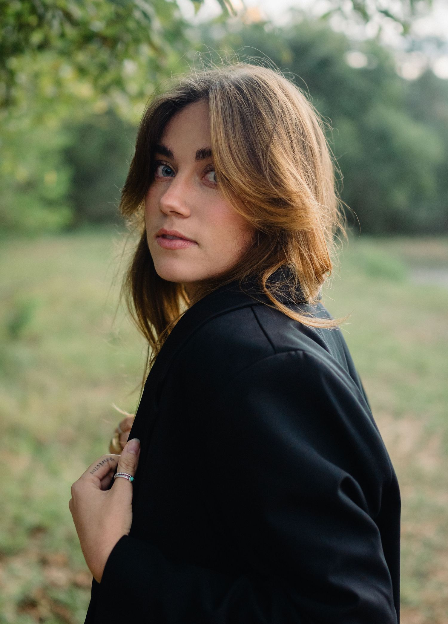 Young woman with brown hair in black jacket glances over shoulder outdoors in green park setting.