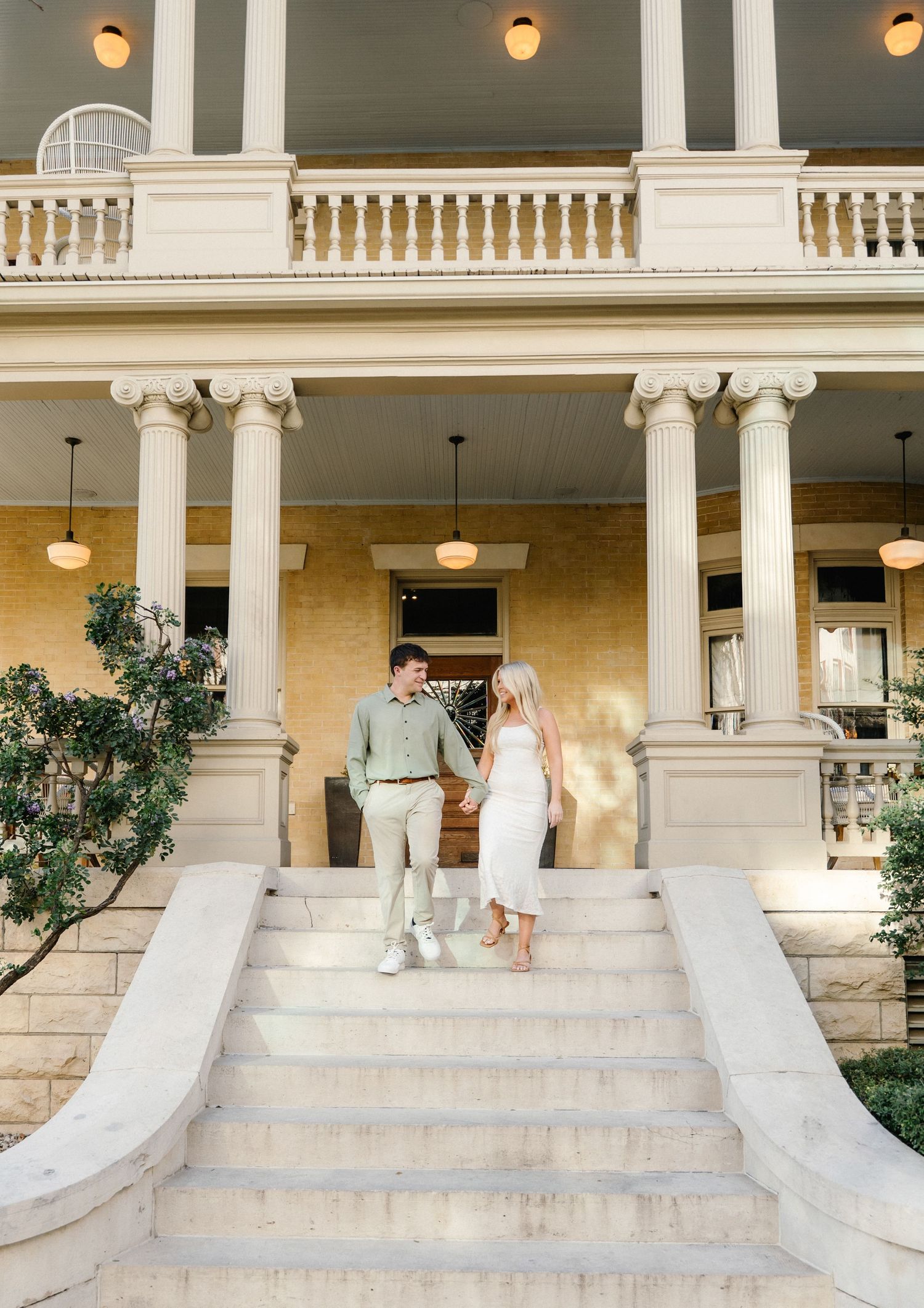 Couple holding hands on grand staircase of elegant historic mansion with white columns and balcony.