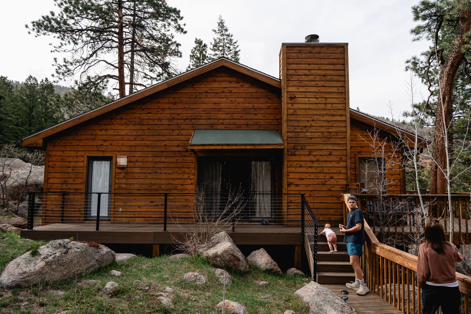 Rustic log cabin with wooden deck and chimney nestled among pine trees and boulders.