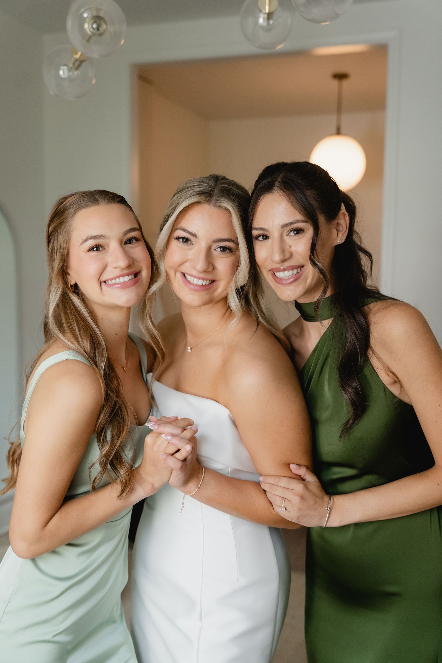 Bride in white dress poses with two bridesmaids in green dresses, smiling together indoors.