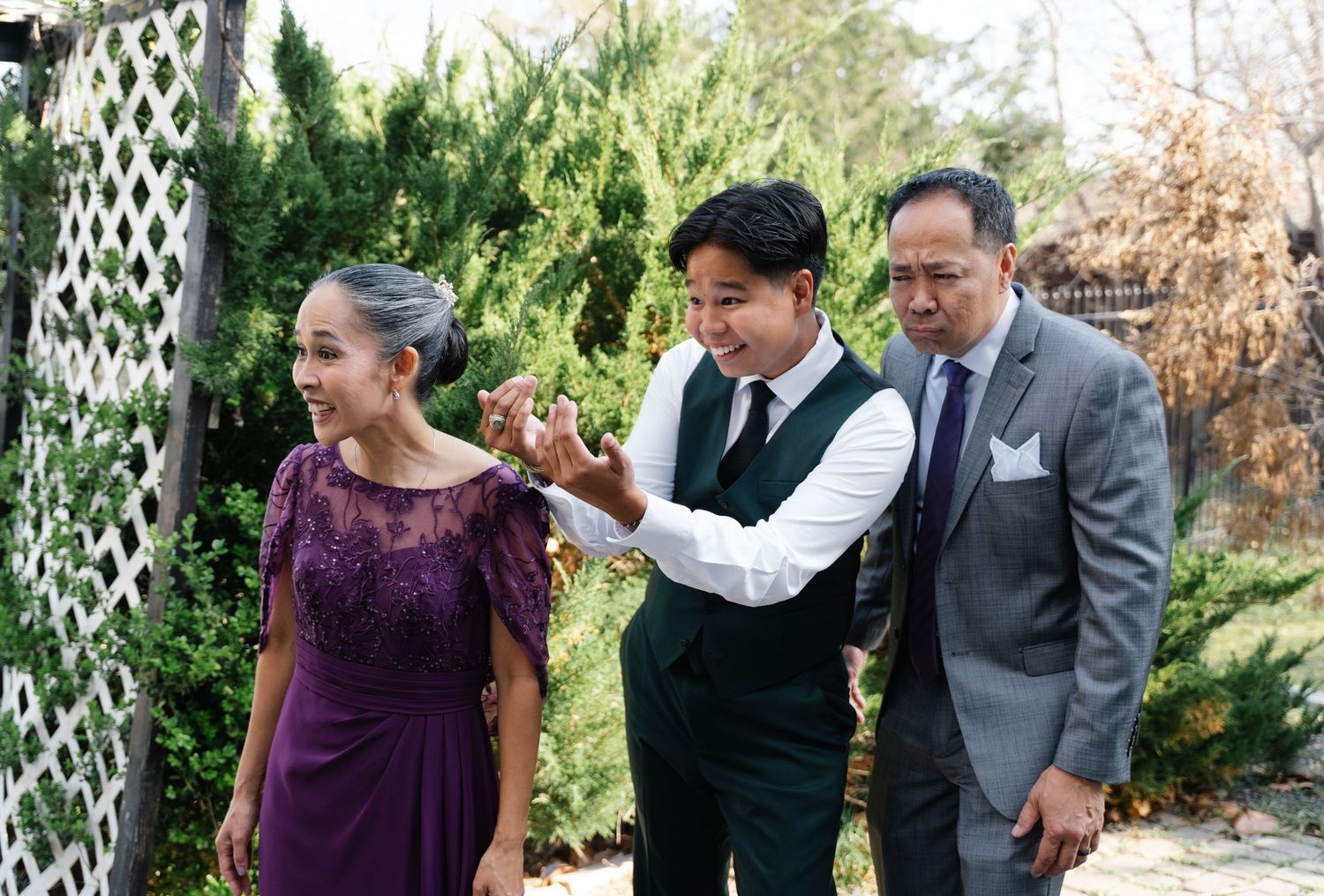 Groom walking with parents outdoors at wedding venue among green trees.