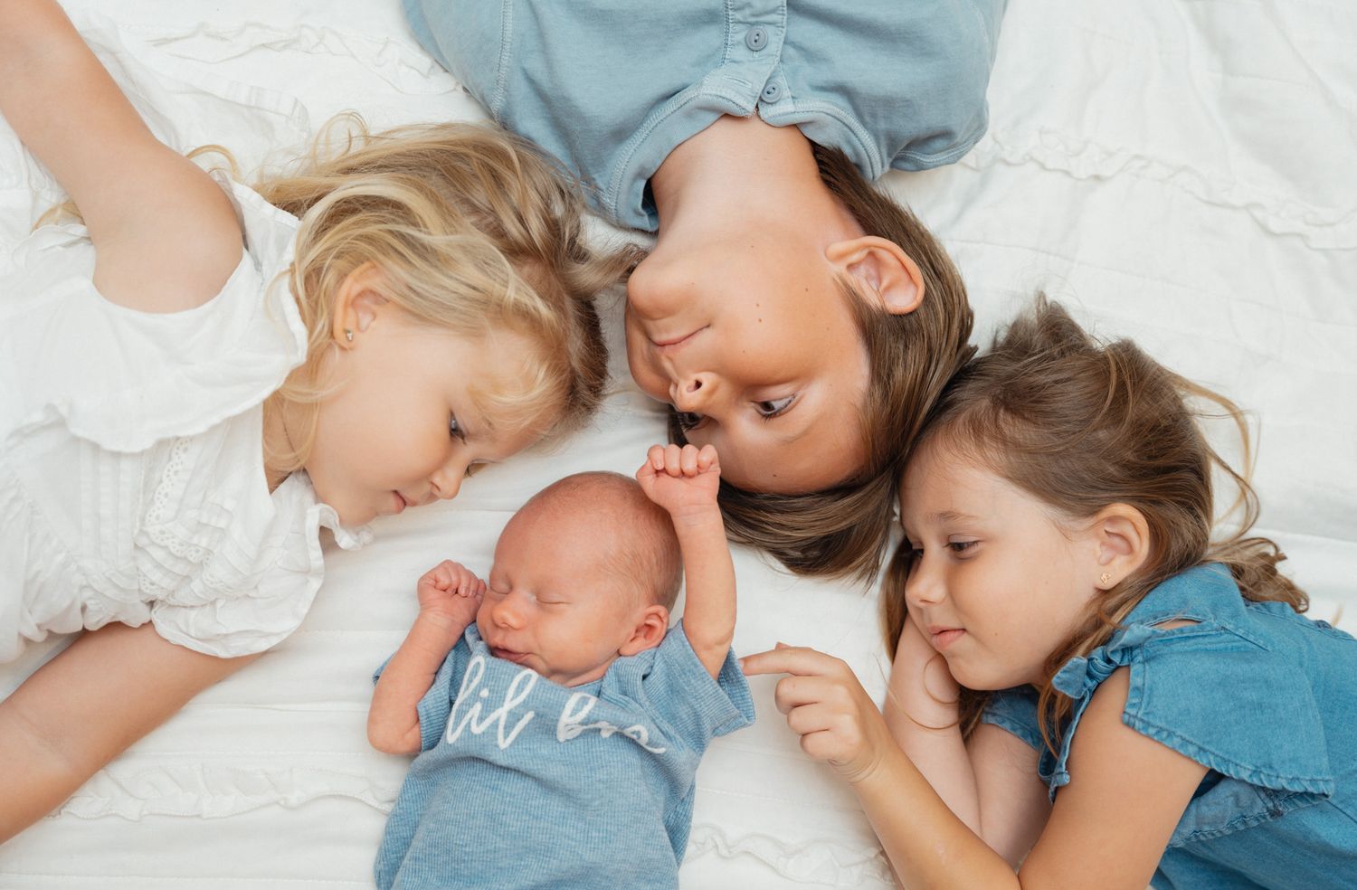 Four children lie on a white bed, lovingly surrounding a peaceful newborn baby.