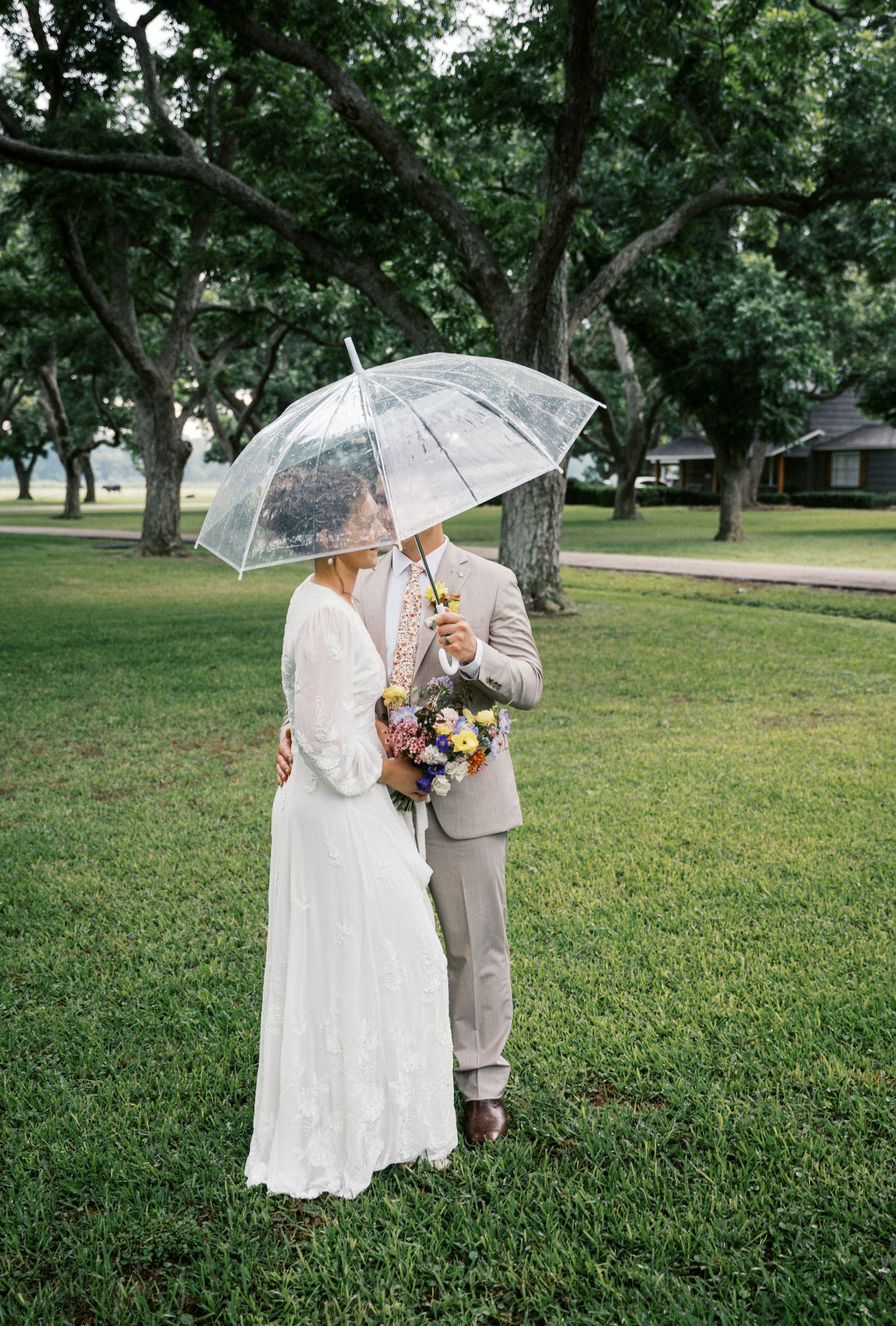 Bride and groom sharing an intimate moment under a clear umbrella on a lush green lawn.