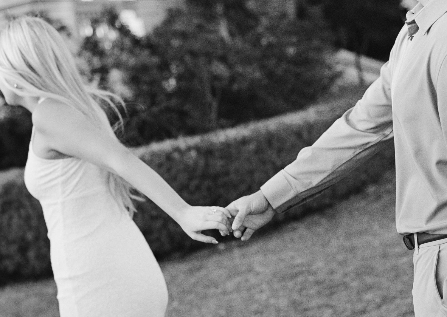 Black and white photo of couple holding hands outdoors, woman in white dress, romantic candid shot.